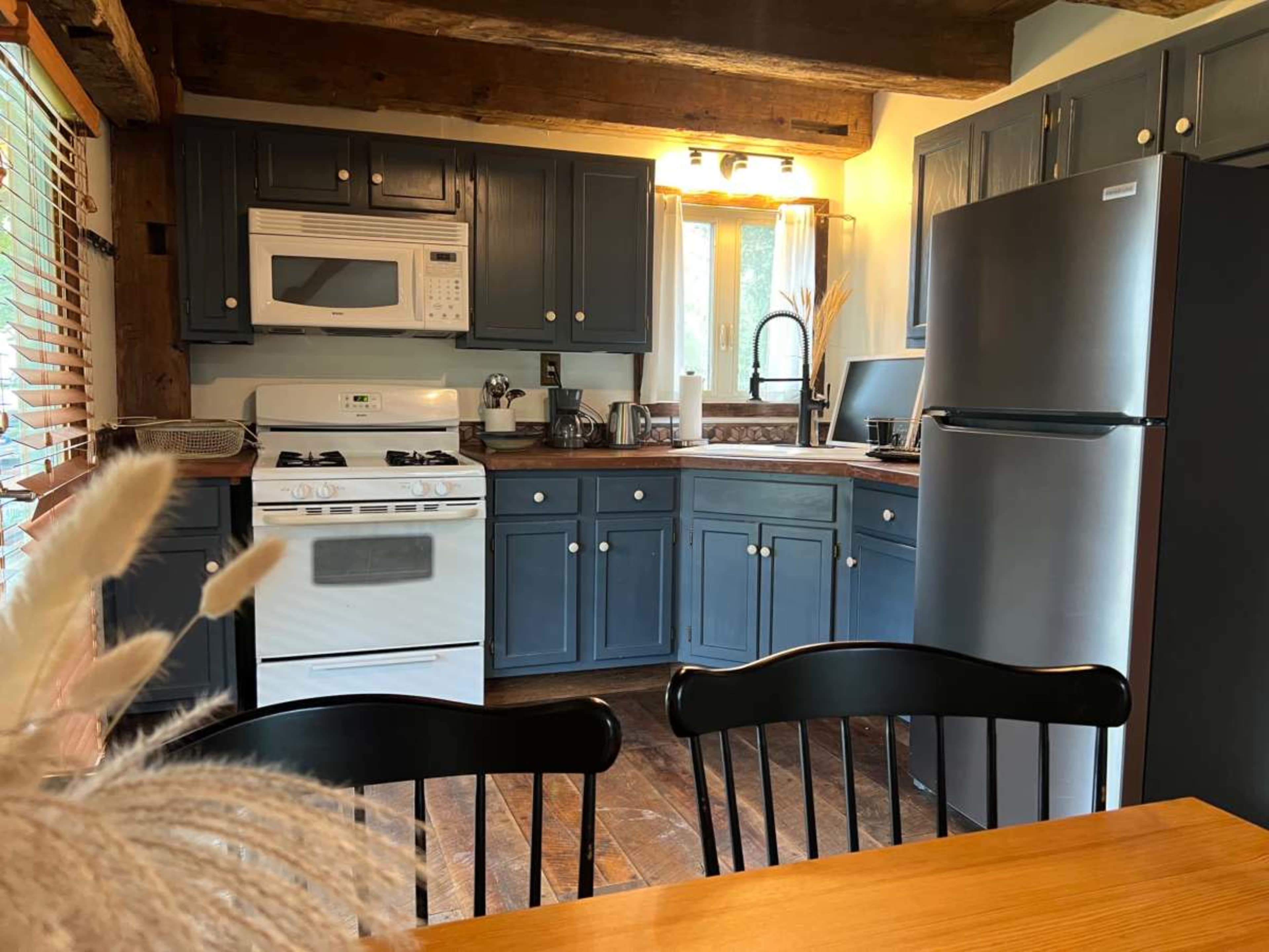 The image shows a cozy kitchen with blue cabinets, a white stove, a microwave, and stainless steel appliances, featuring a wooden dining table in the foreground.