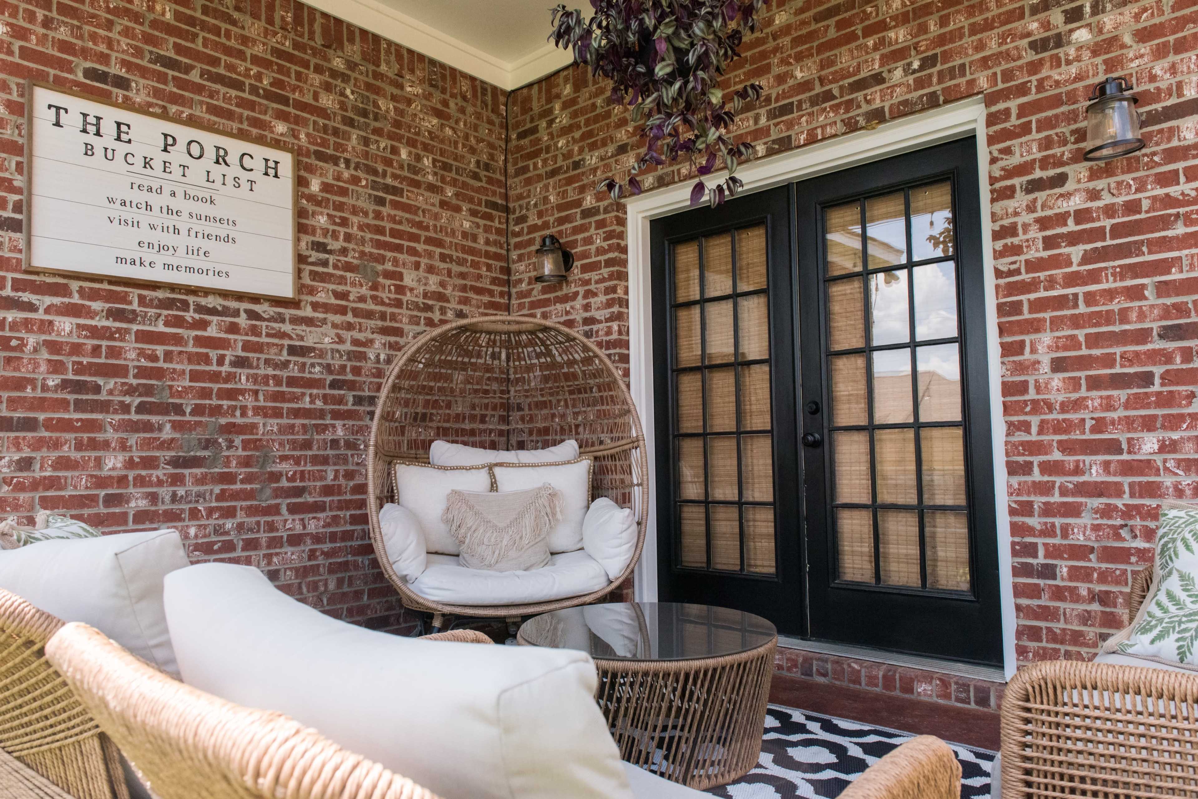 A cozy porch area featuring a hanging rattan chair, a round glass table, and a decorative sign on the brick wall.