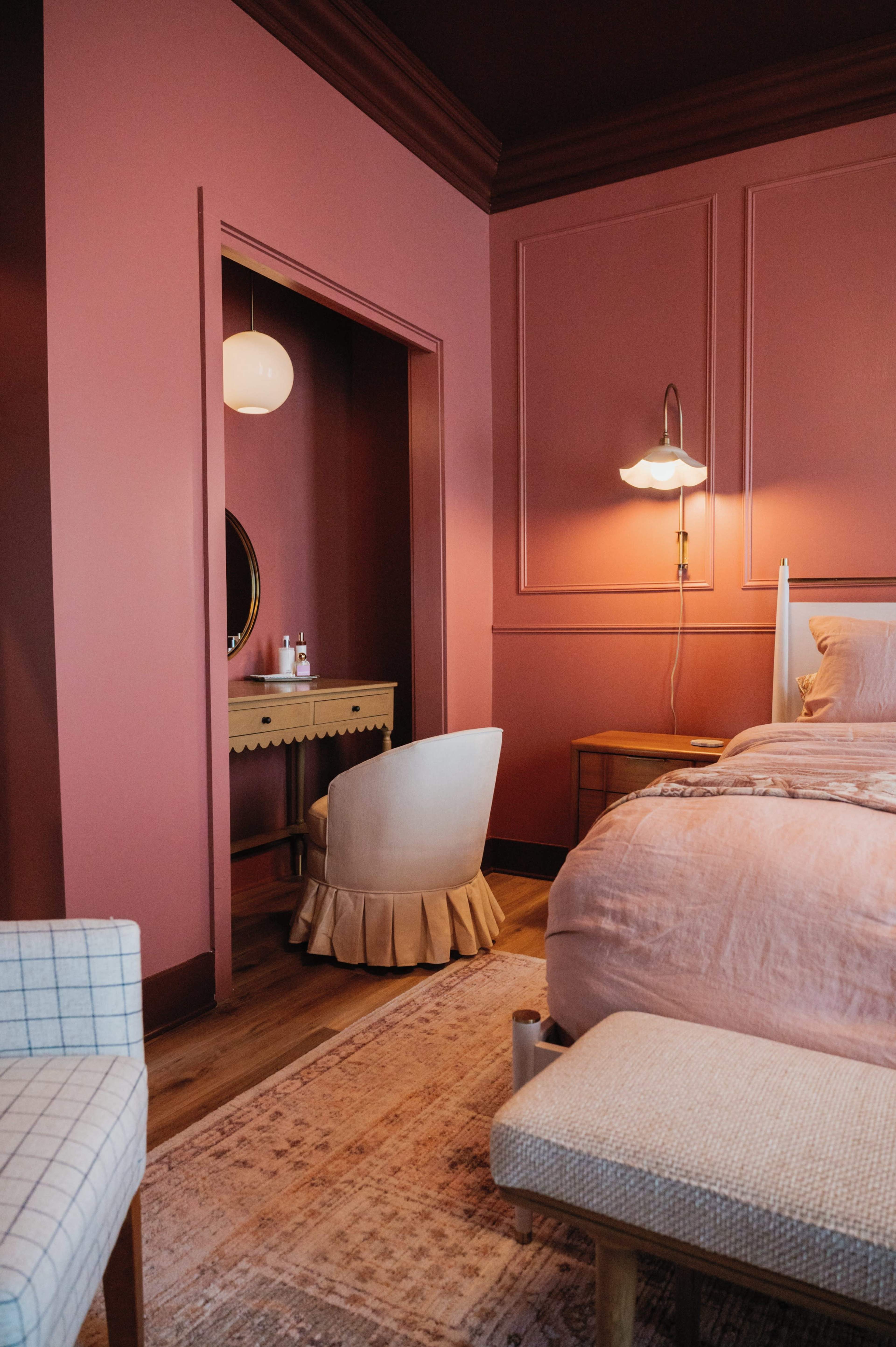 The image shows a stylish bedroom with a pink feature wall, a wooden bed, a small desk with a circular mirror, and a decorative chair.