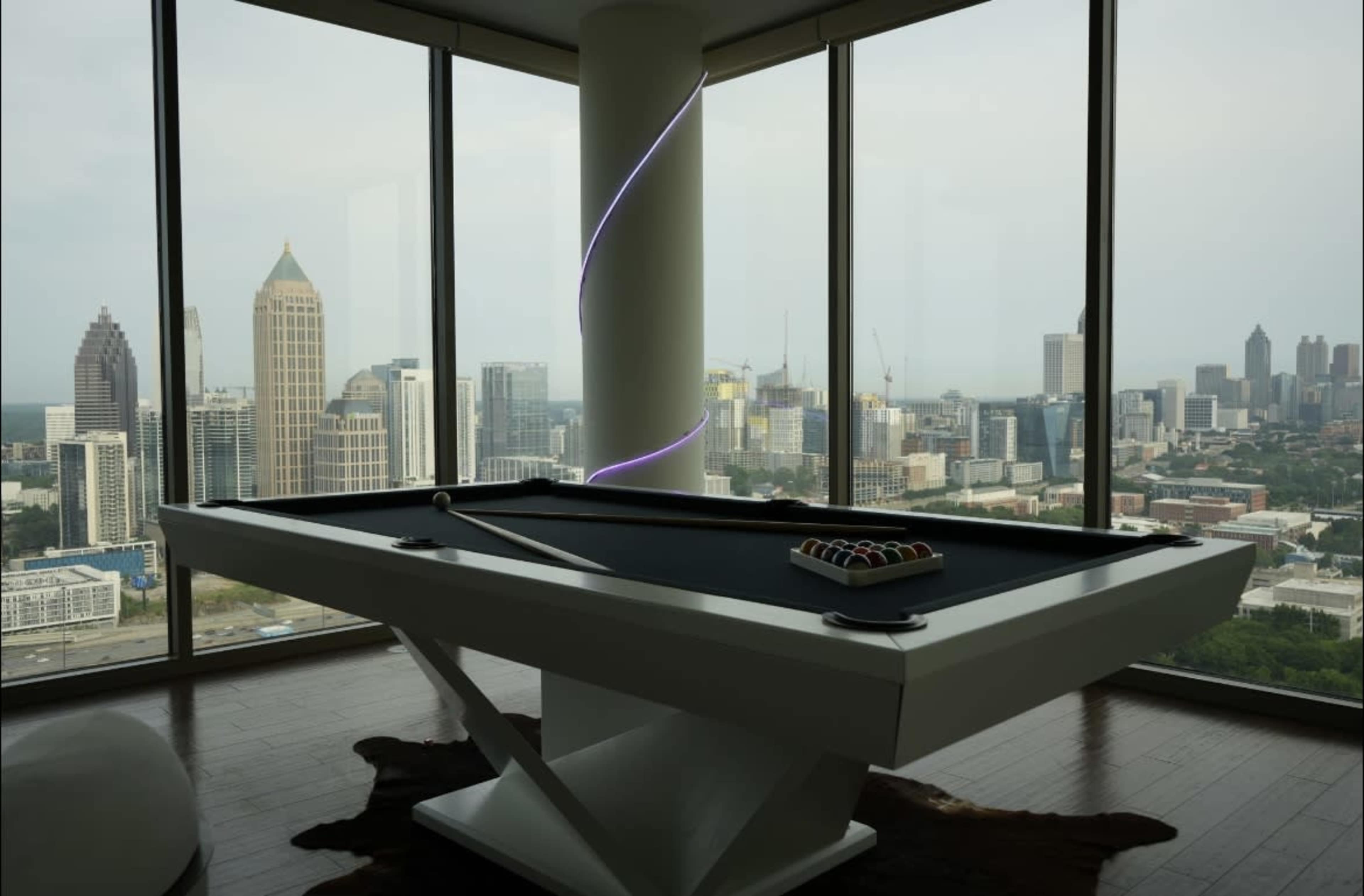 An elevated pool table is positioned in a modern room with large windows overlooking a city skyline.