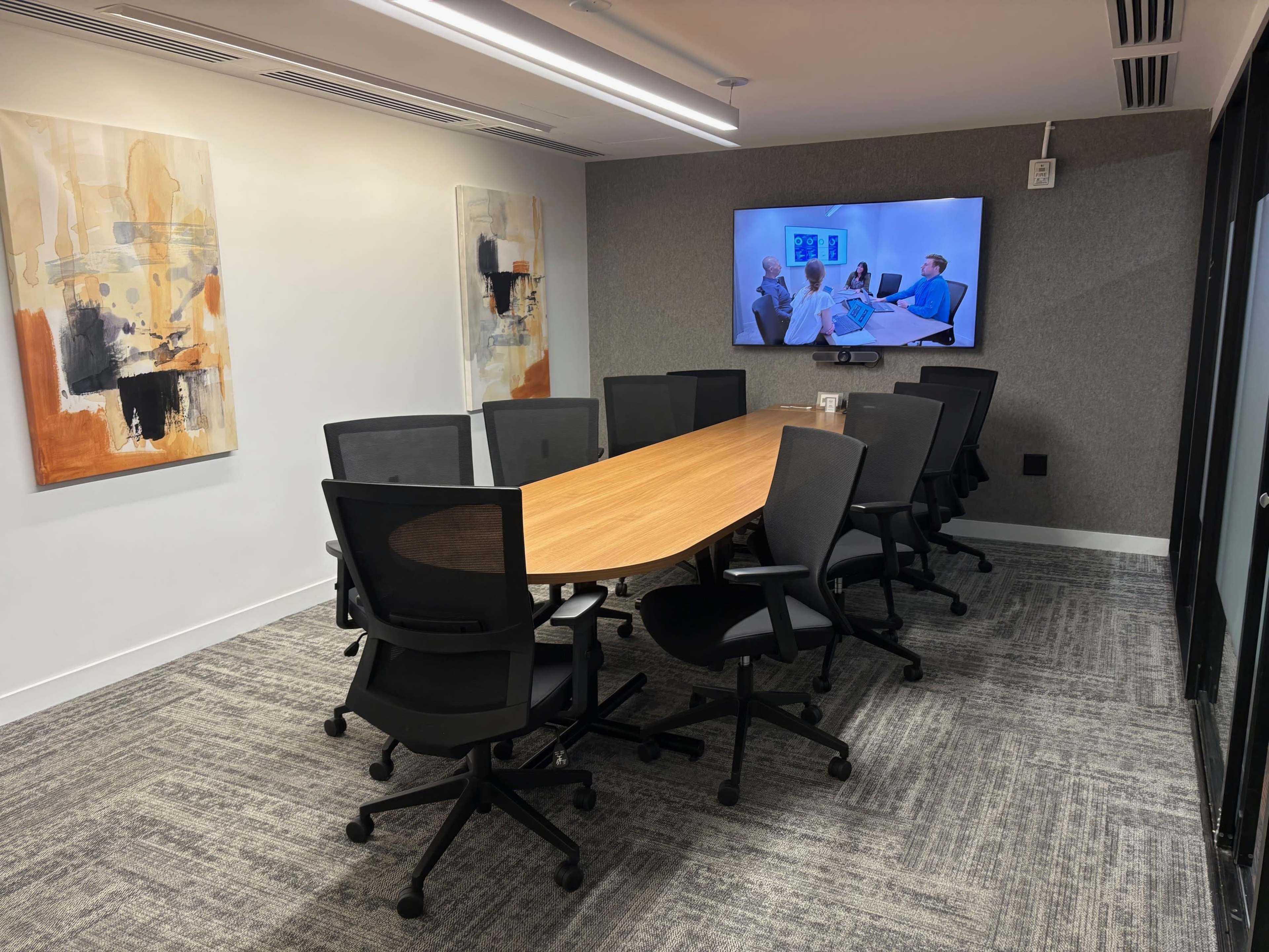 A modern conference room features a long wooden table surrounded by black mesh chairs, with two abstract paintings on the wall and a television mounted on the opposite wall.