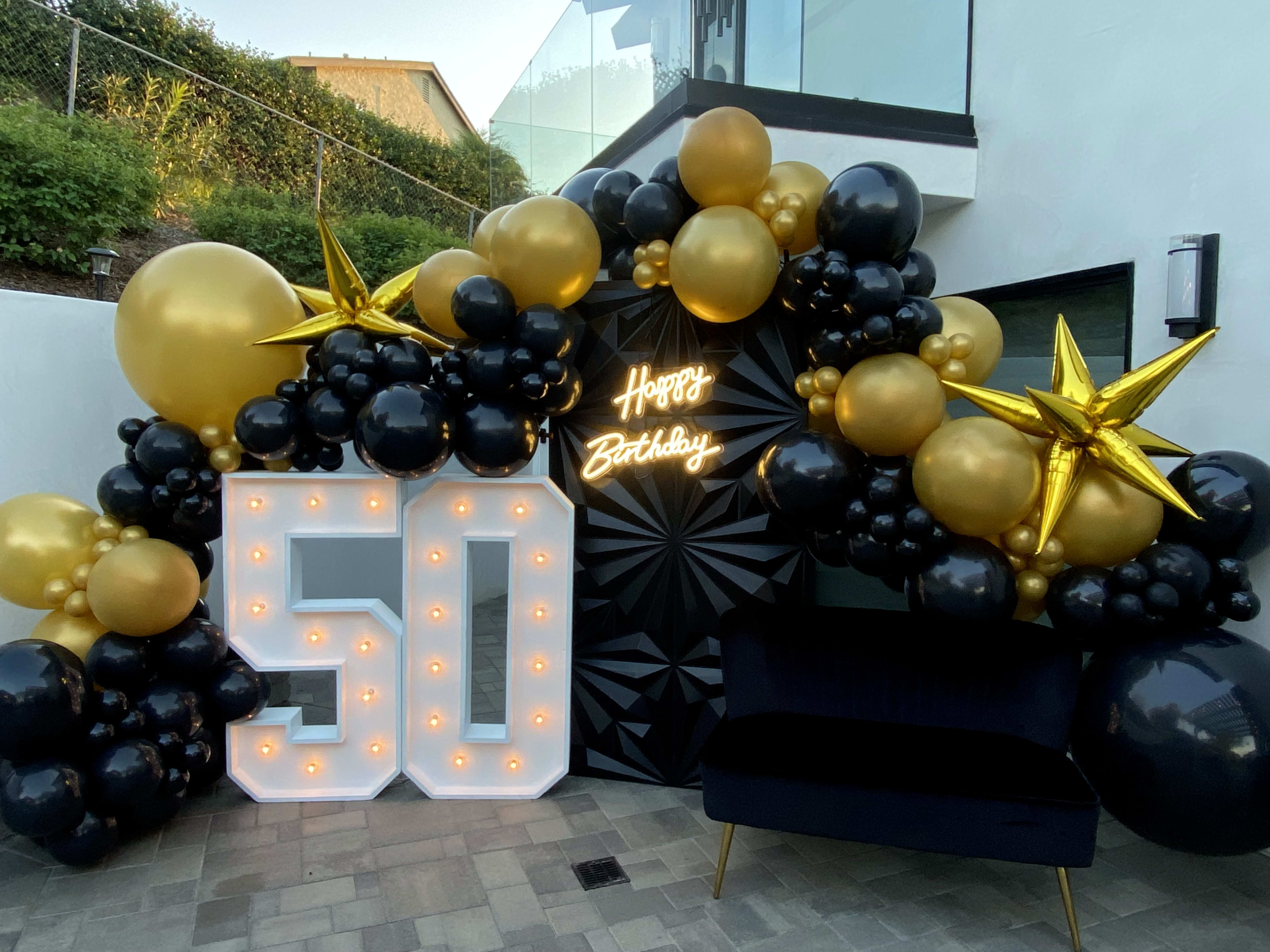 The scene features a 50th birthday celebration setup with large illuminated numbers "50," a "Happy Birthday" neon sign, and an arch of black, gold, and balloon decorations.