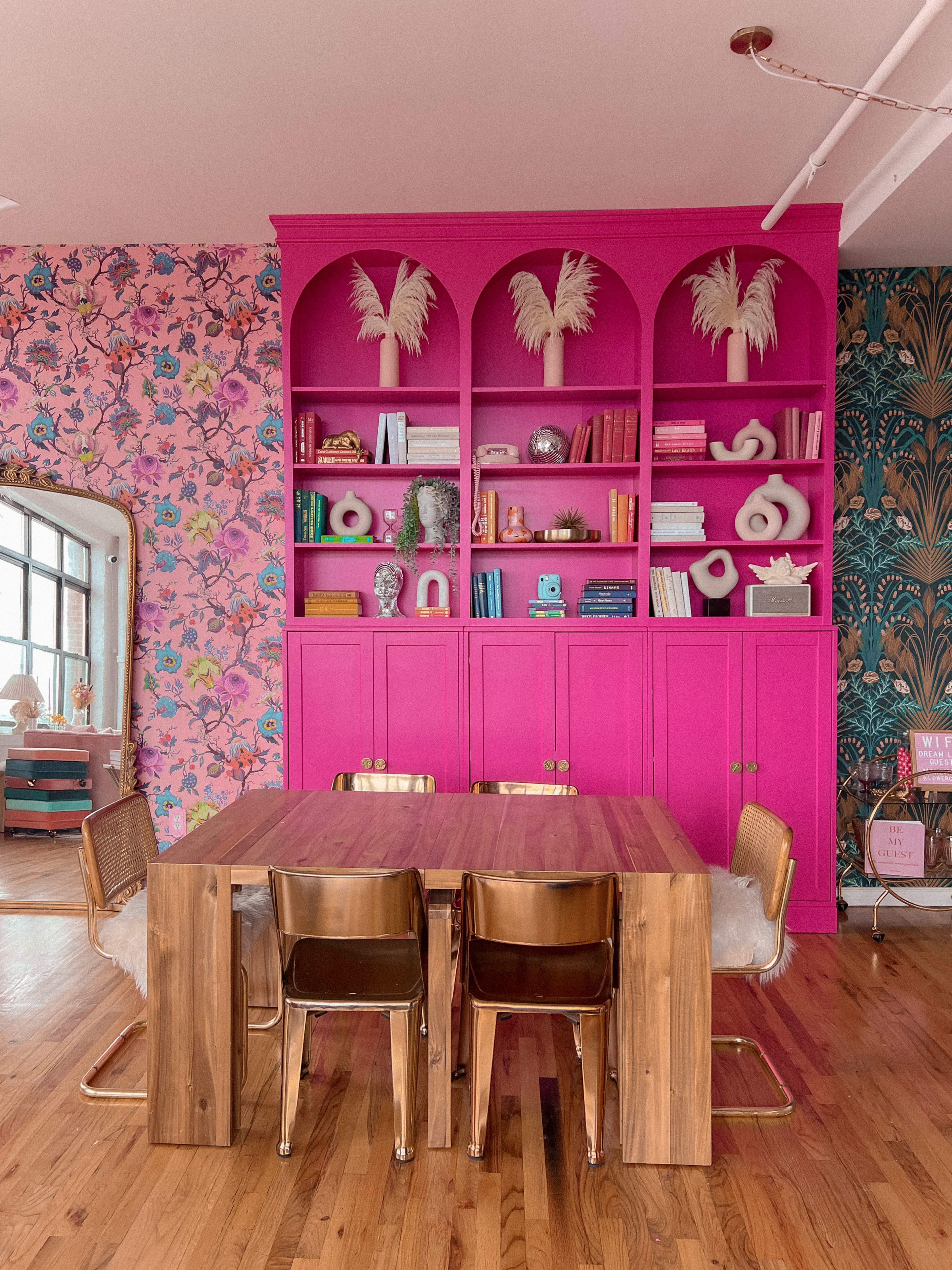 A colorful dining area features a large wooden table surrounded by metallic chairs, with a vibrant pink bookshelf filled with decorative items and books in the background.