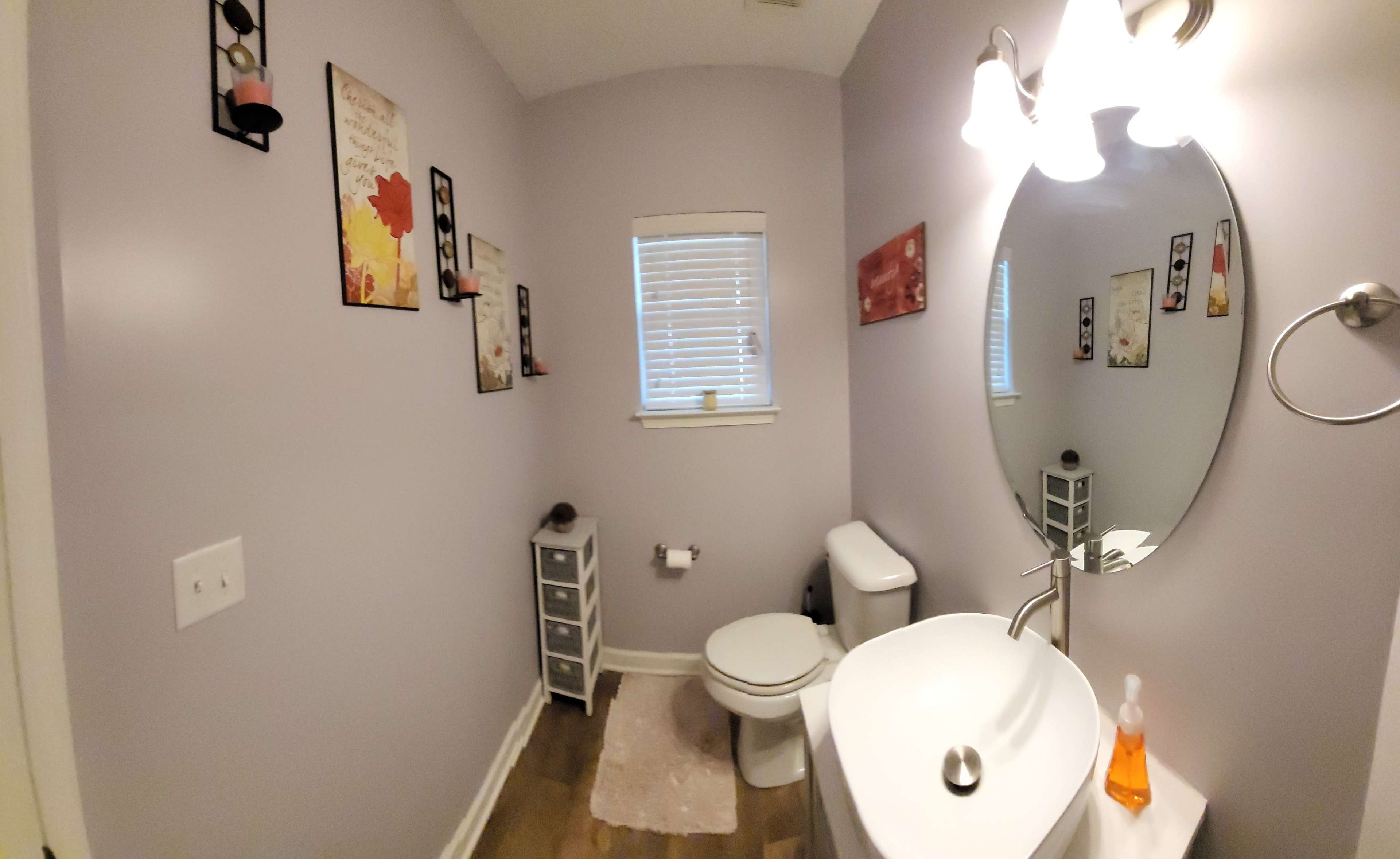 A small bathroom with light purple walls, a round mirror above a sink, a toilet, a small storage cabinet, and decorative wall art.