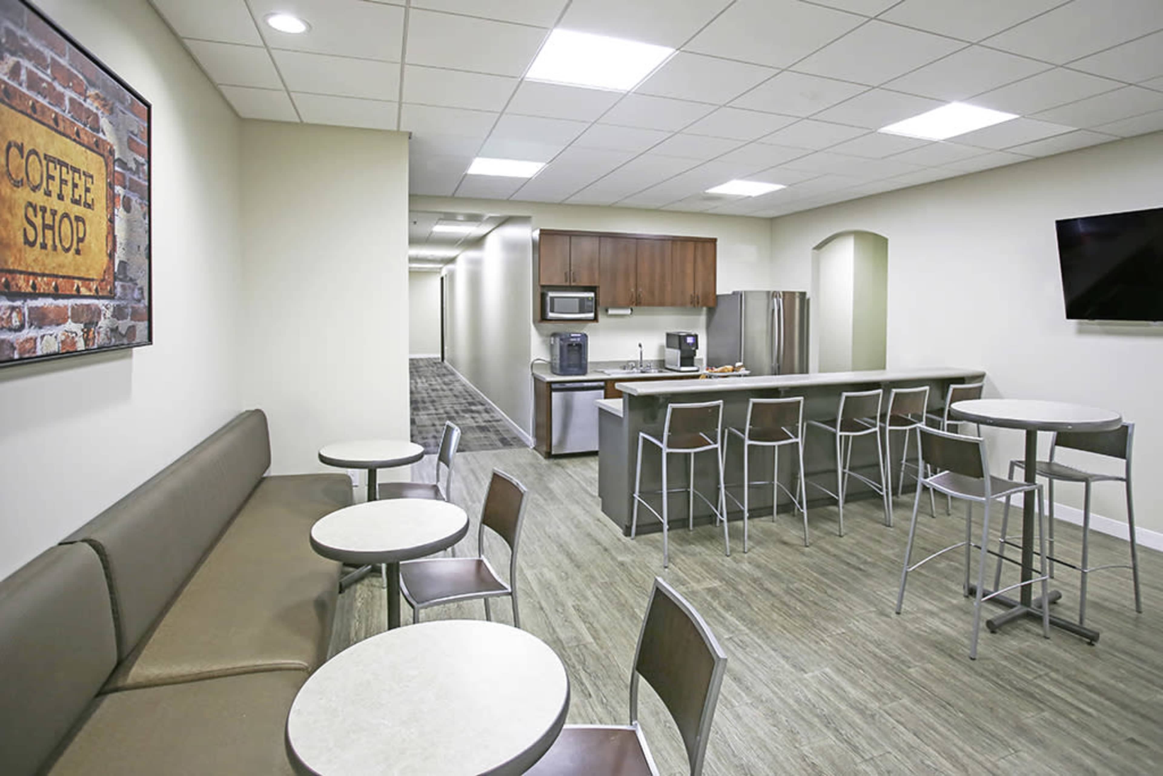 A modern break room with seating, a coffee shop sign, and kitchen appliances along one wall.