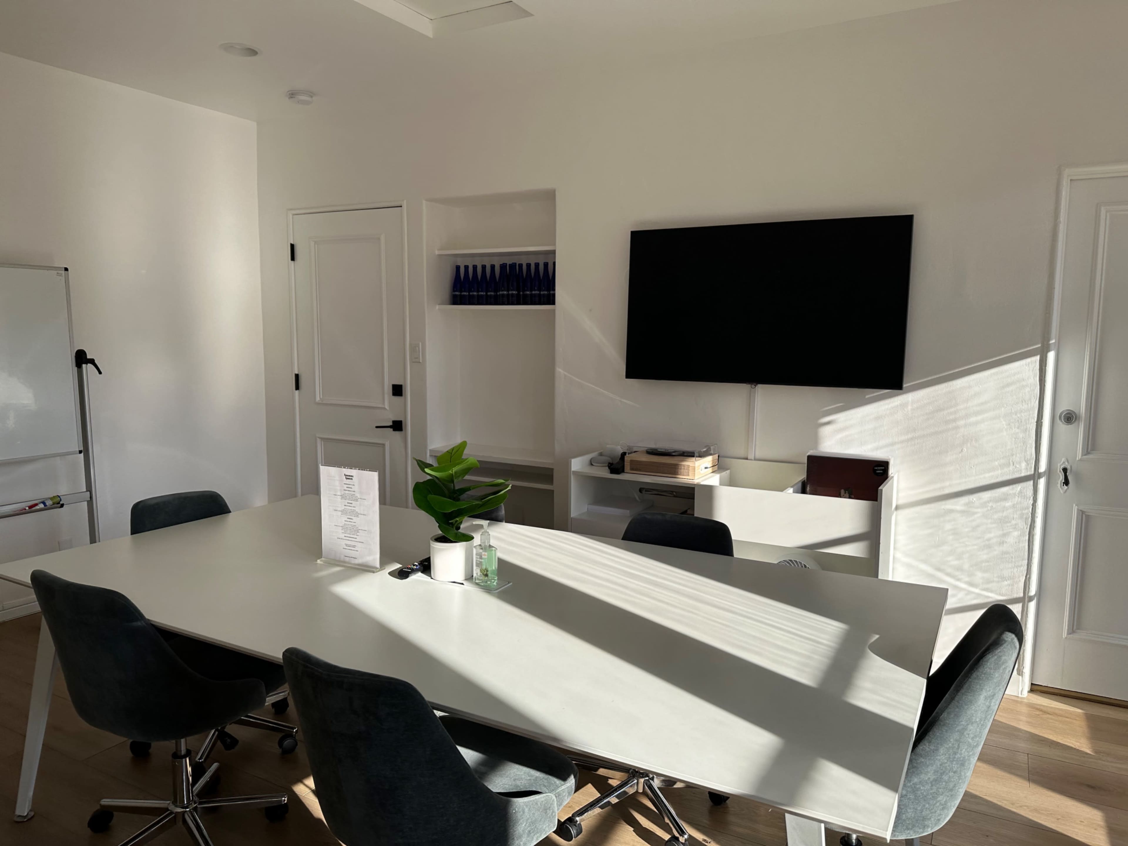 A modern meeting room featuring a large table, ergonomic chairs, a wall-mounted TV, and a plant on the table, with sunlight casting shadows across the space.