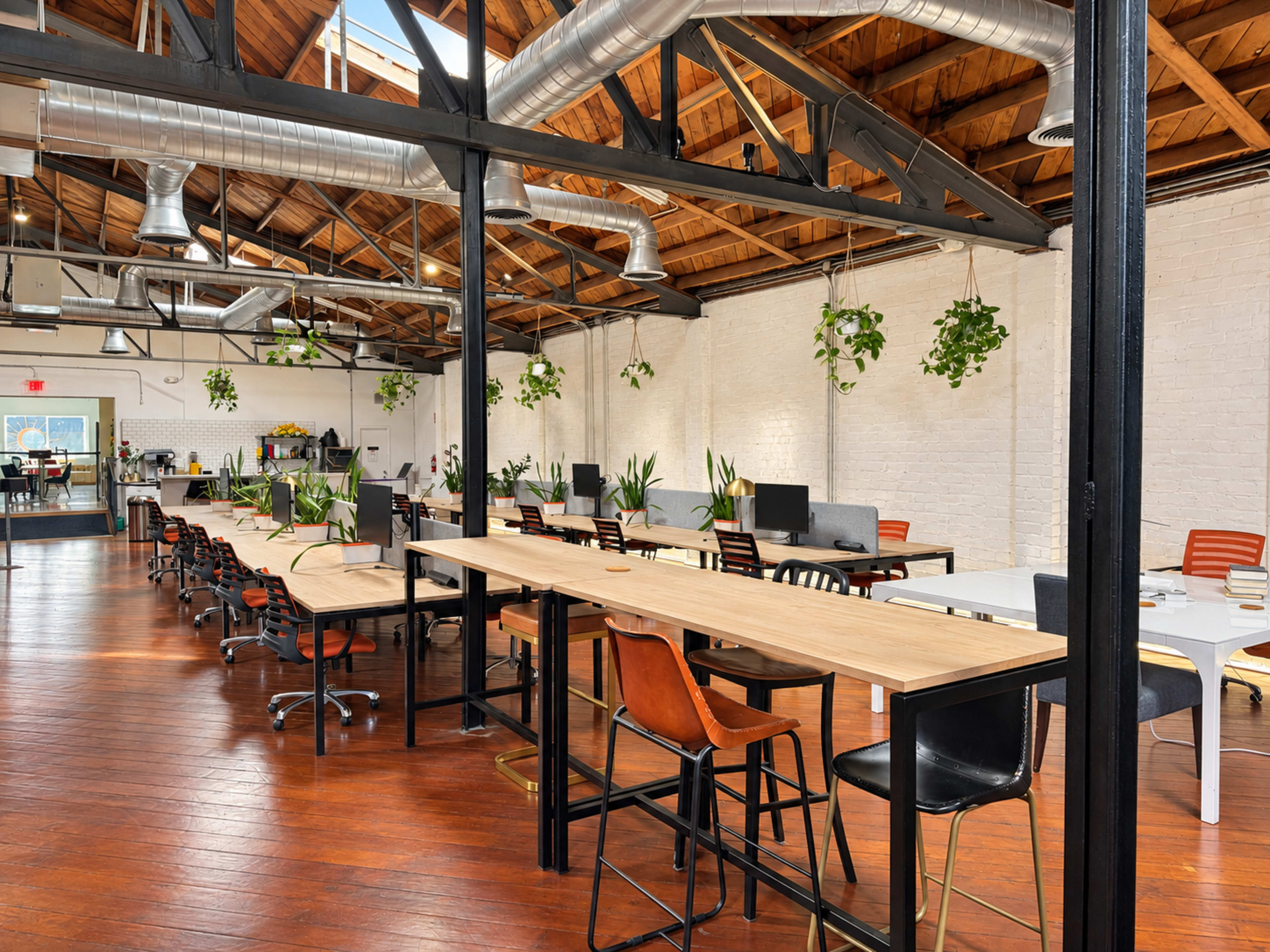 The image shows a spacious, modern office interior with long tables, office chairs, and greenery hanging from the ceiling.