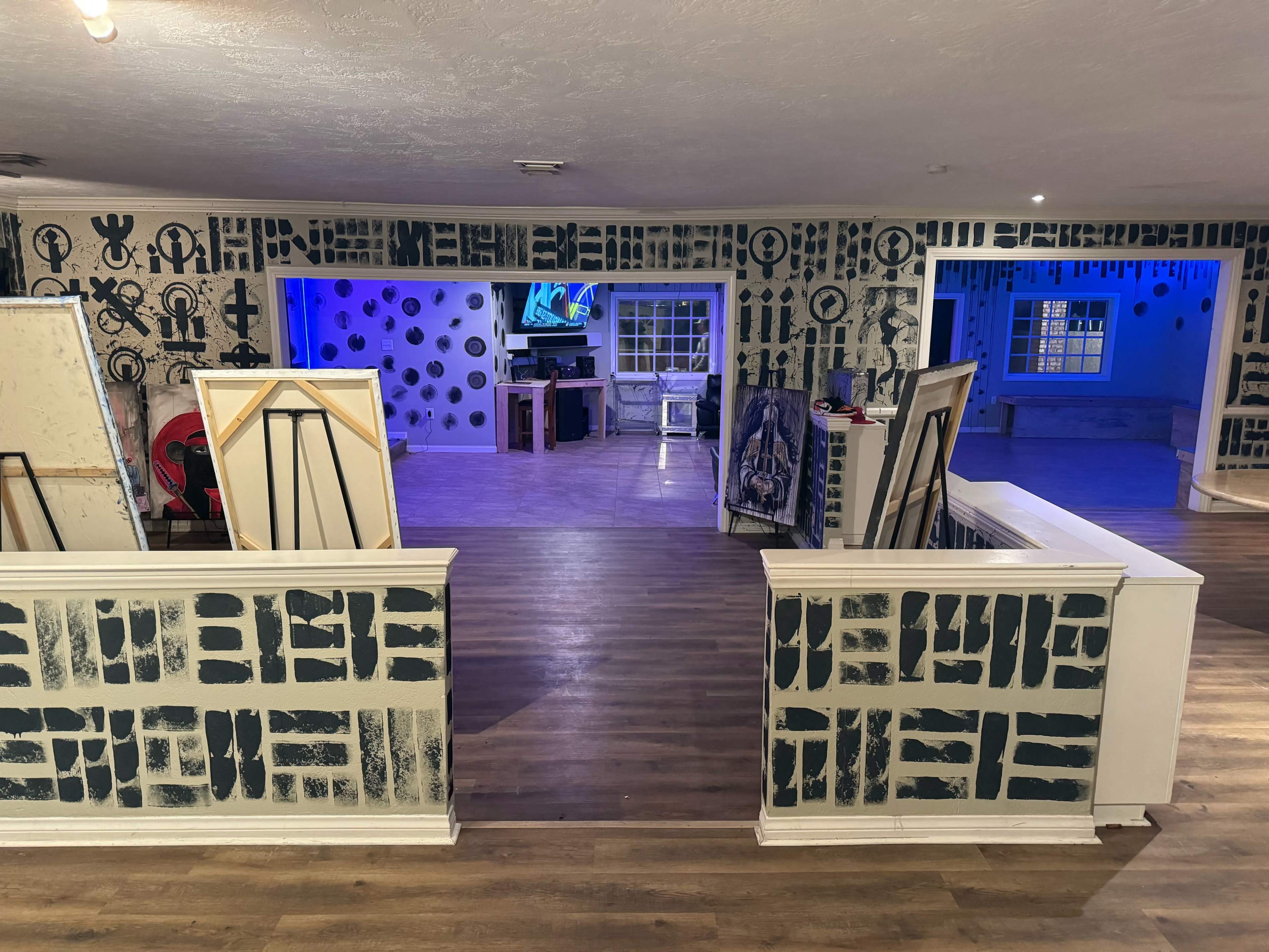 A spacious interior with patterned walls and multiple framed artworks leaning against the railing, leading to an area with blue lighting and a television.