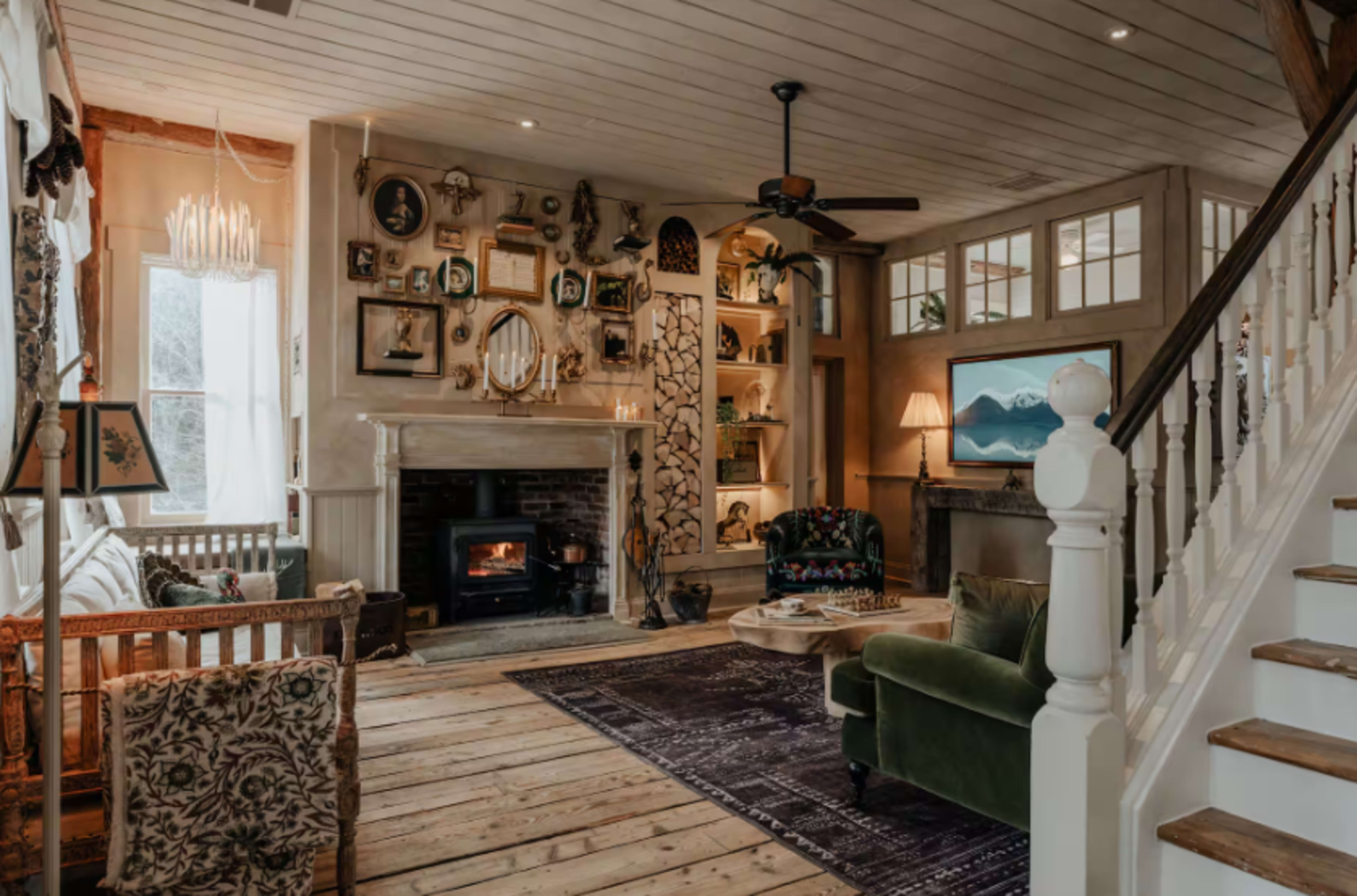 The image shows a cozy living room with a fireplace, wooden flooring, and walls adorned with various framed pictures and decorative items.