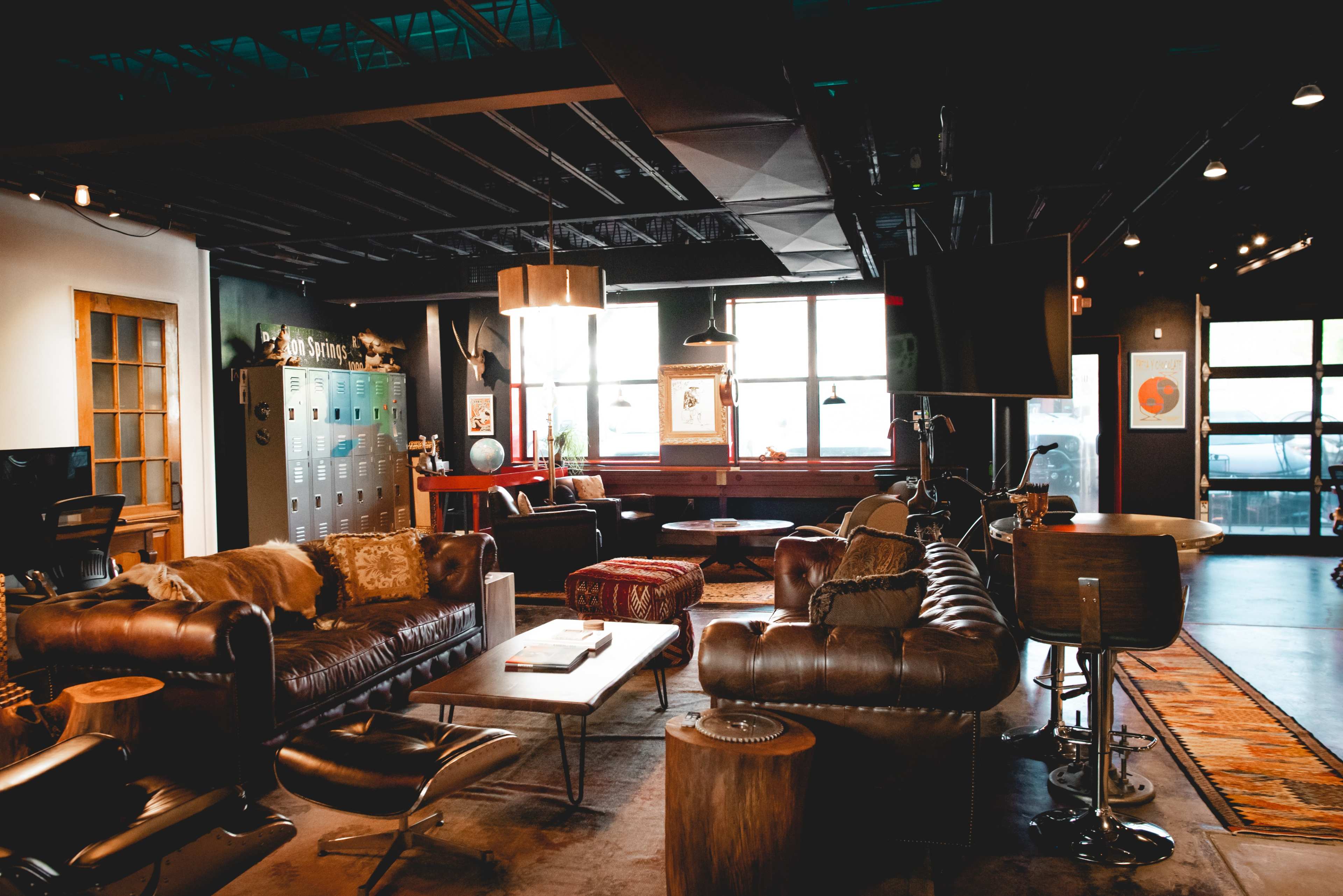 The image shows a modern lounge area featuring leather sofas, wooden tables, and large windows allowing natural light to enter.