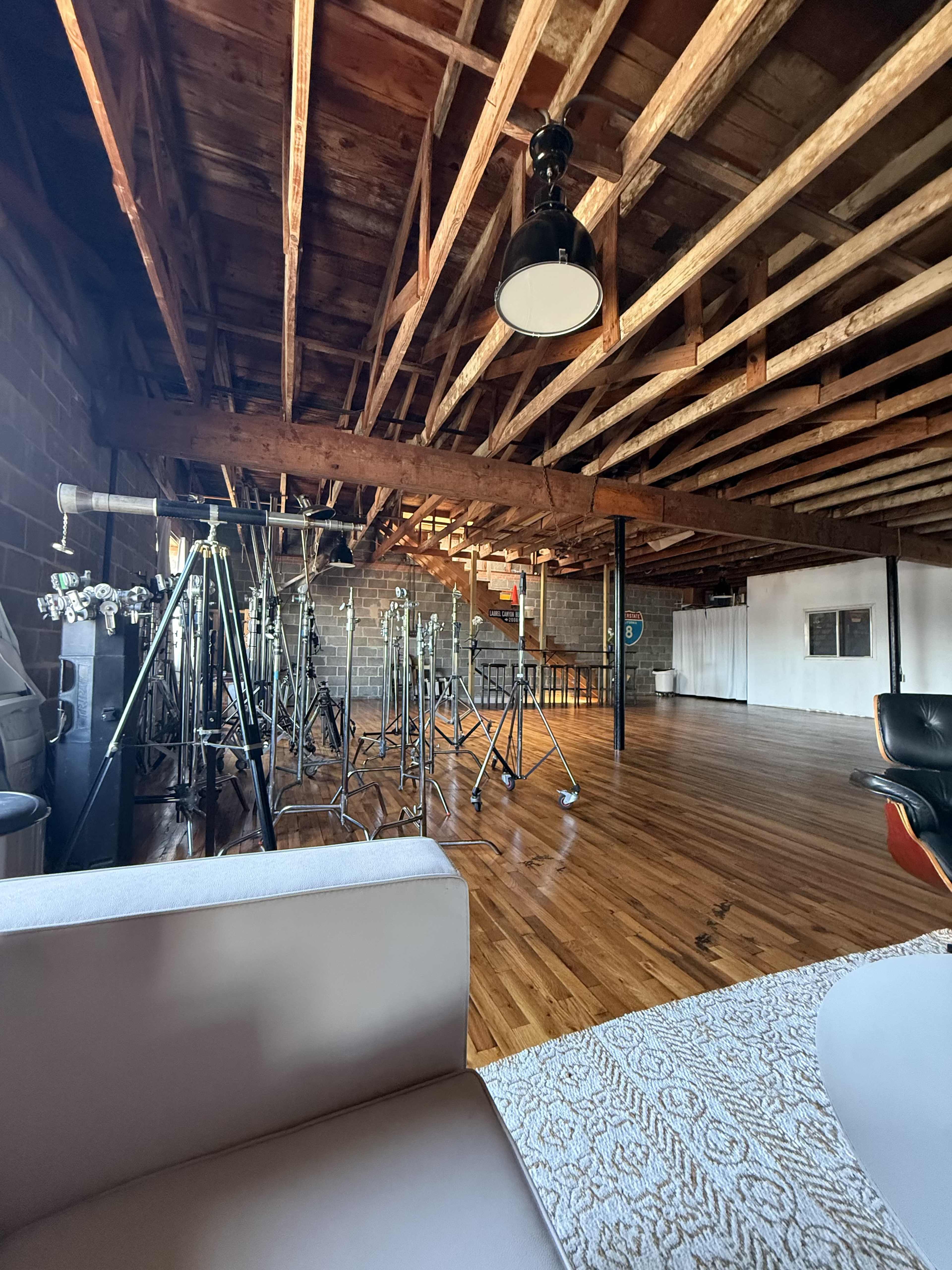 The image shows a spacious room with exposed wooden beams on the ceiling, featuring metal stands in the background and a polished wooden floor.