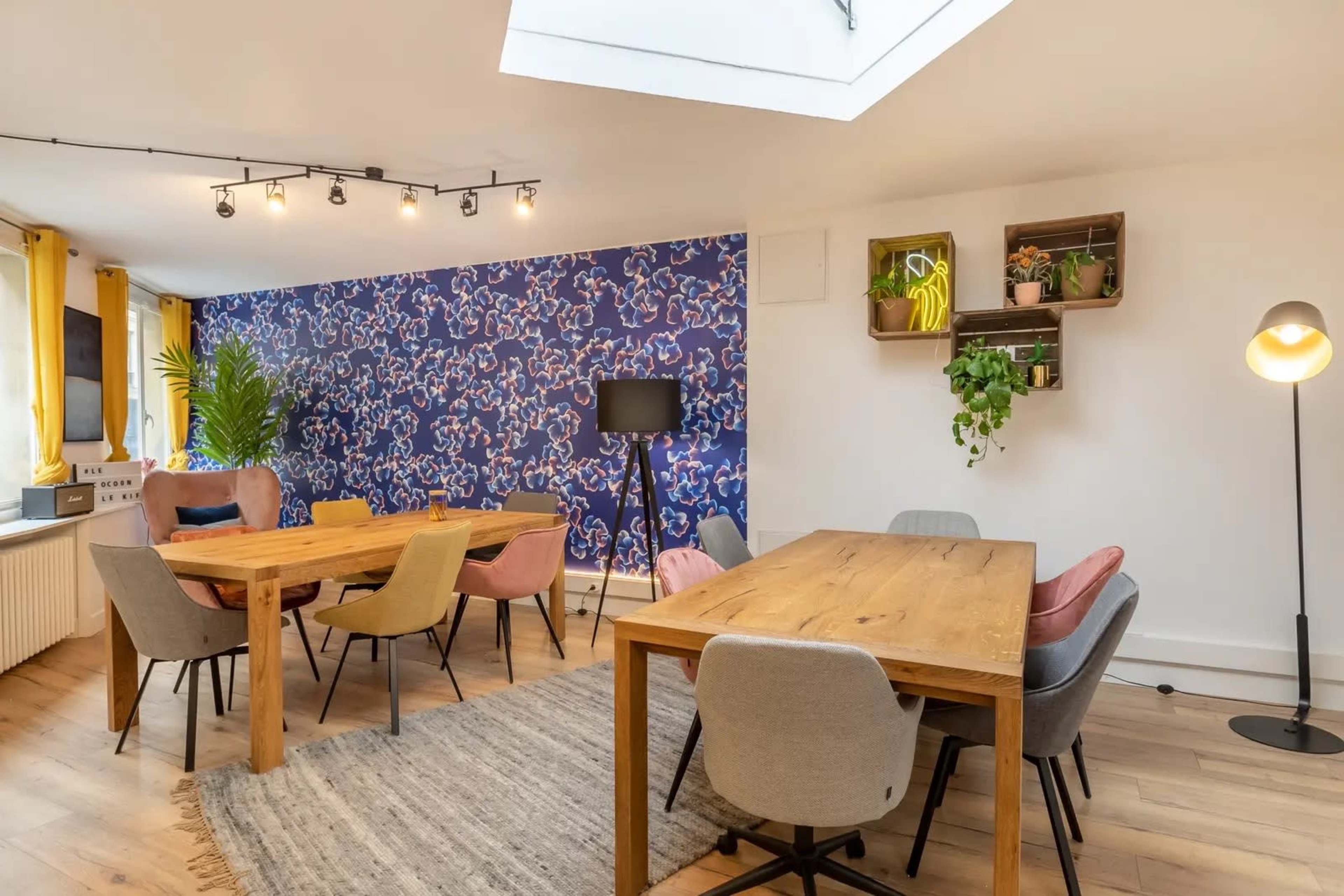 A brightly lit dining area features wooden tables surrounded by upholstered chairs, with a patterned blue wall and plants enhancing the decor.