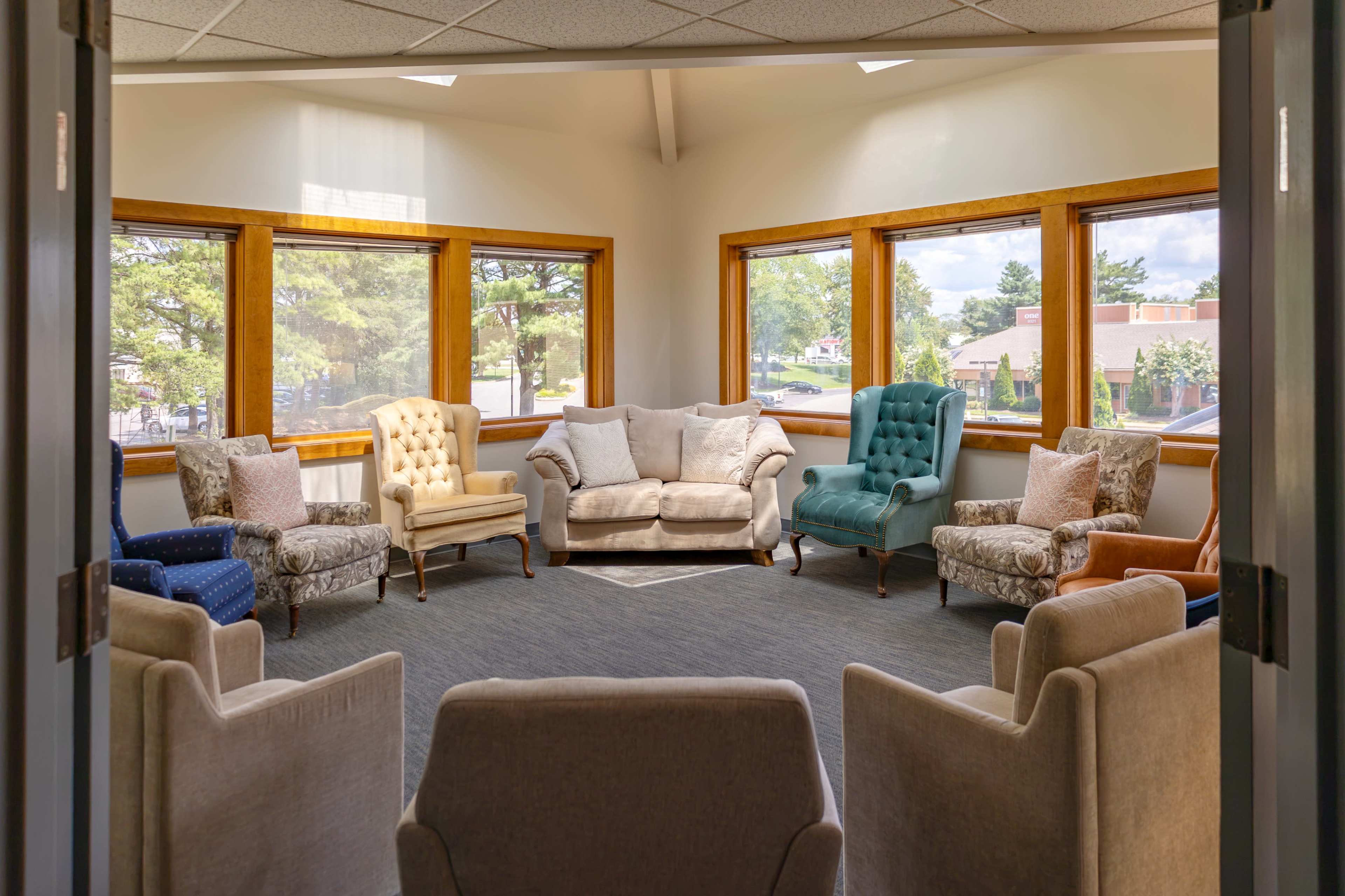 The image shows a seating area with various upholstered chairs arranged around a central space, surrounded by windows that let in natural light.