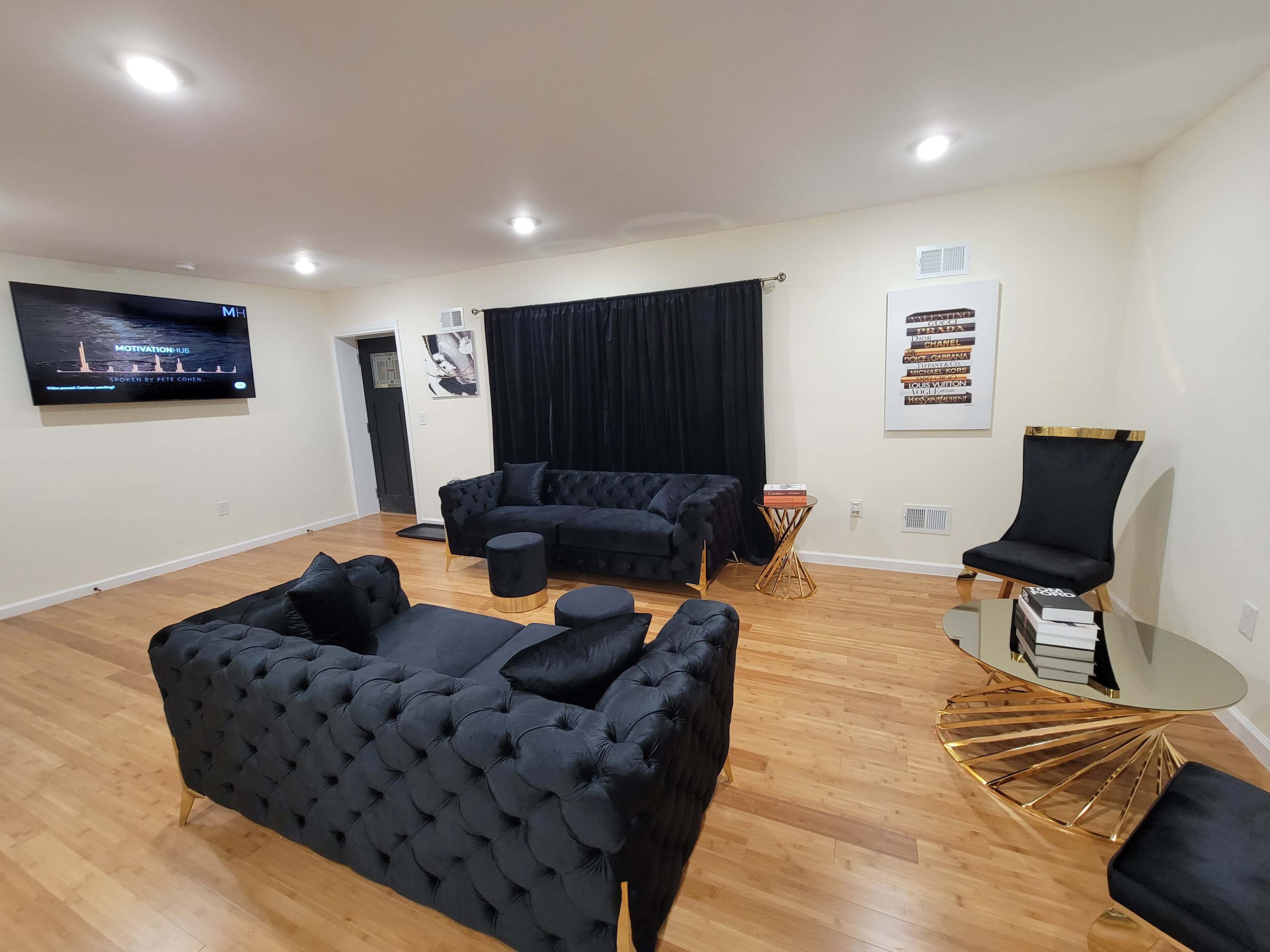The room features black tufted furniture, a large flat-screen TV on the wall, and a round gold accent table with books.