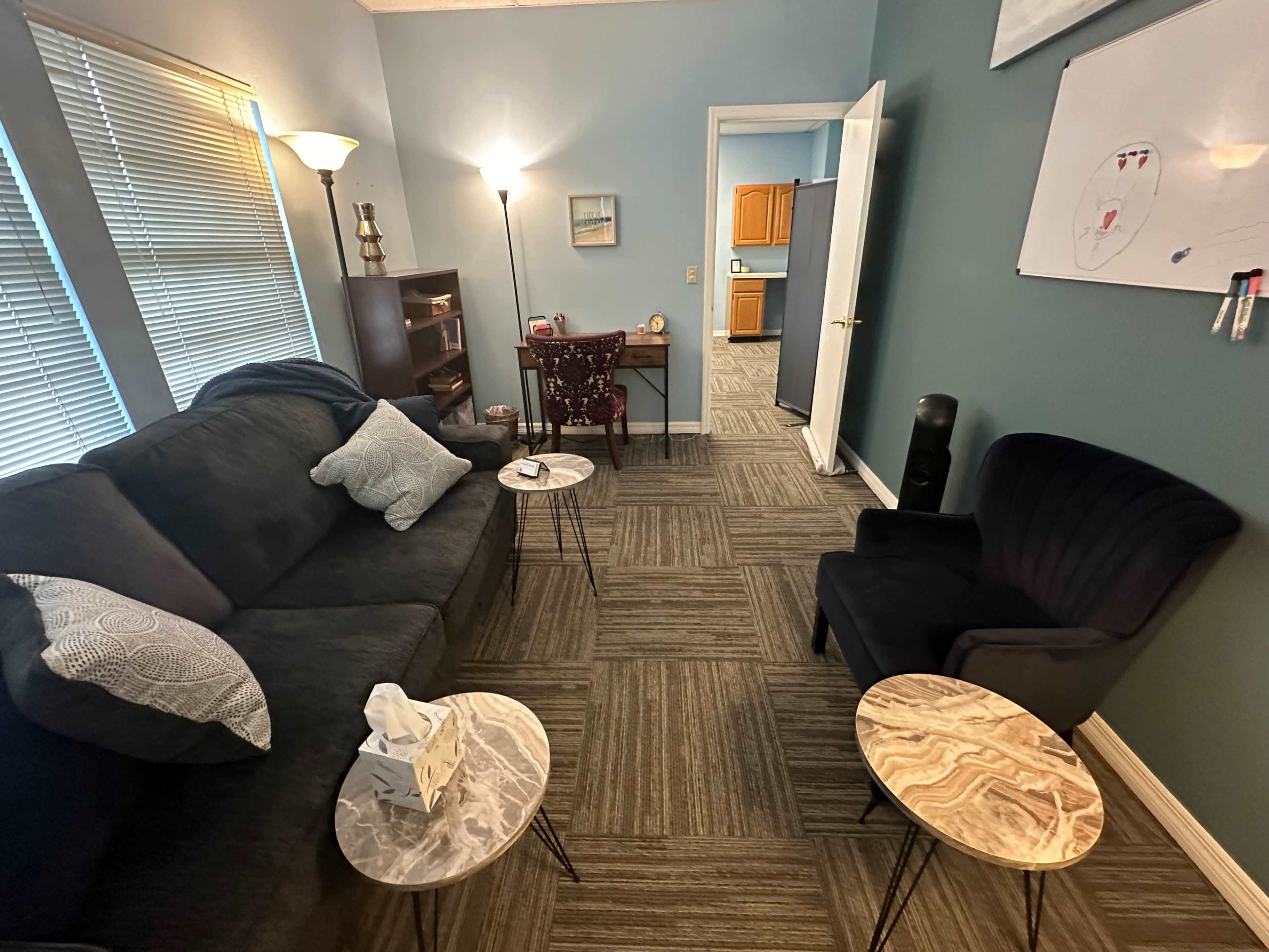 The image shows a cozy room with a dark sofa, two small marble tables, a chair, and a door leading to a kitchen area.