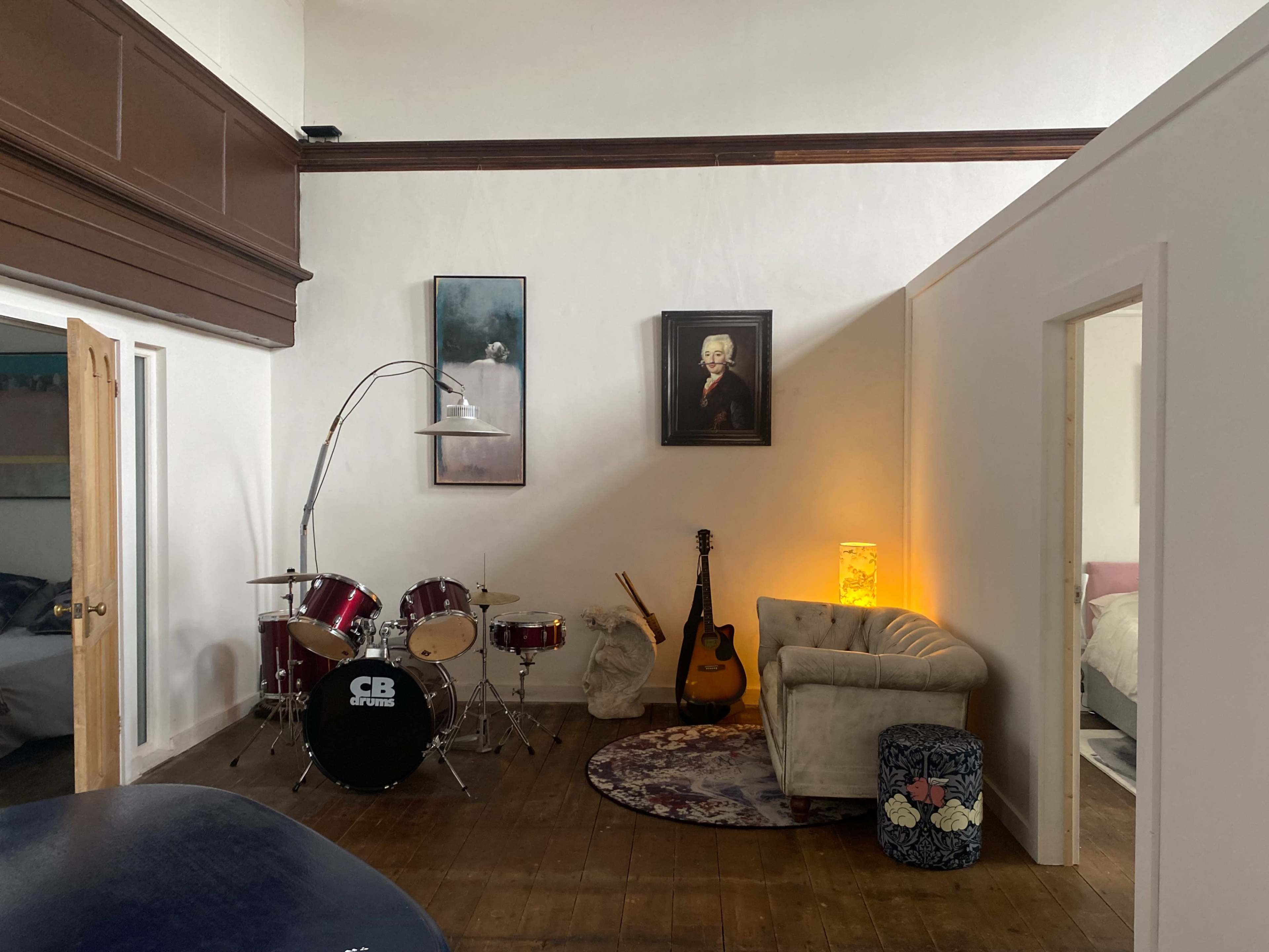 A cozy room with a drum set, an acoustic guitar, and a vintage sofa, illuminated by warm light from a lamp.