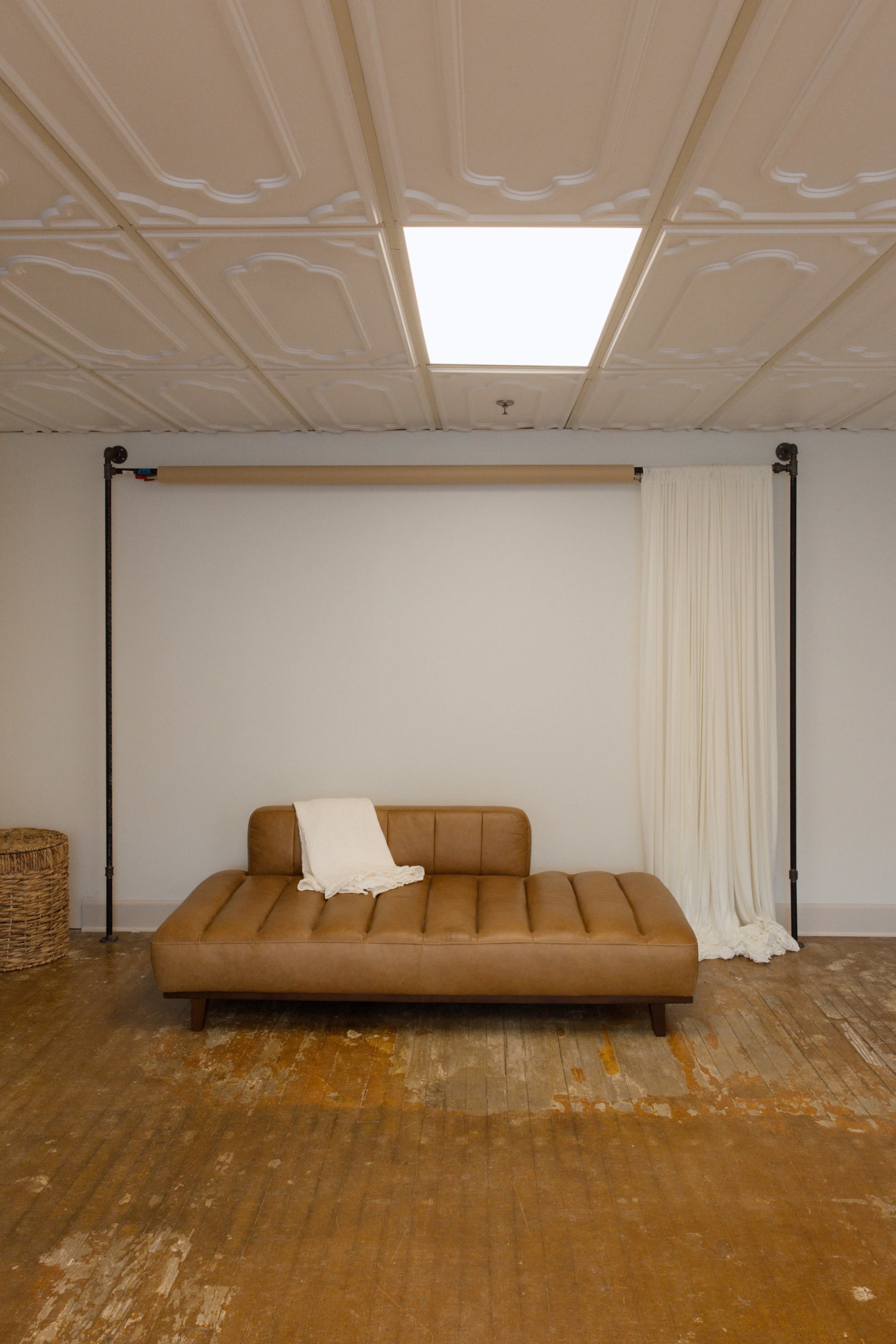 The image shows a minimalist room with a brown upholstered sofa, a white fabric draping from a metal rod, and a woven basket on a wooden floor.
