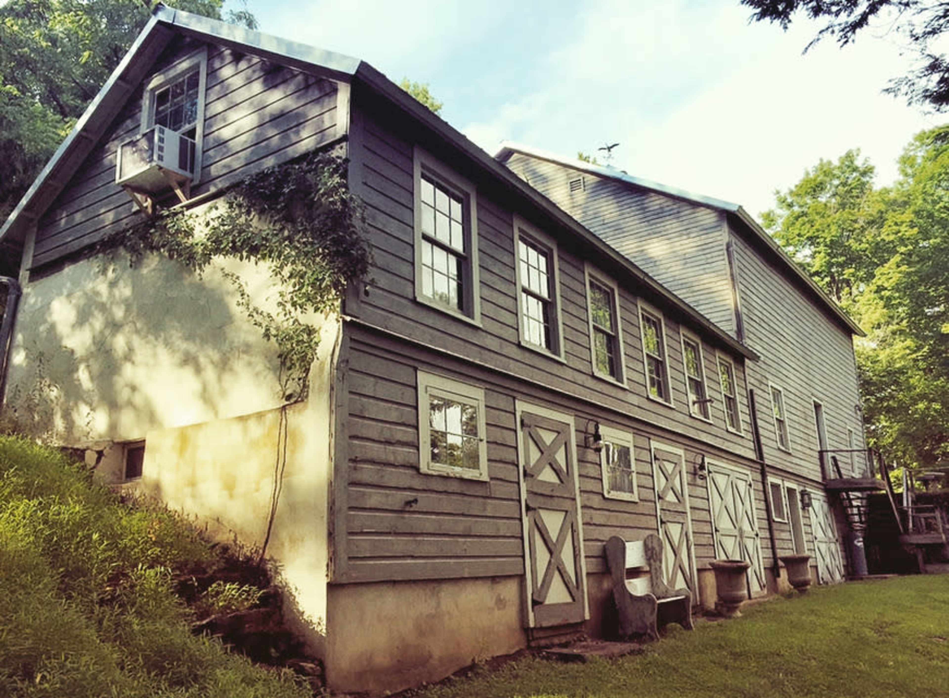 A weathered wooden barn with multiple windows and doors, situated on a grassy hillside surrounded by trees.