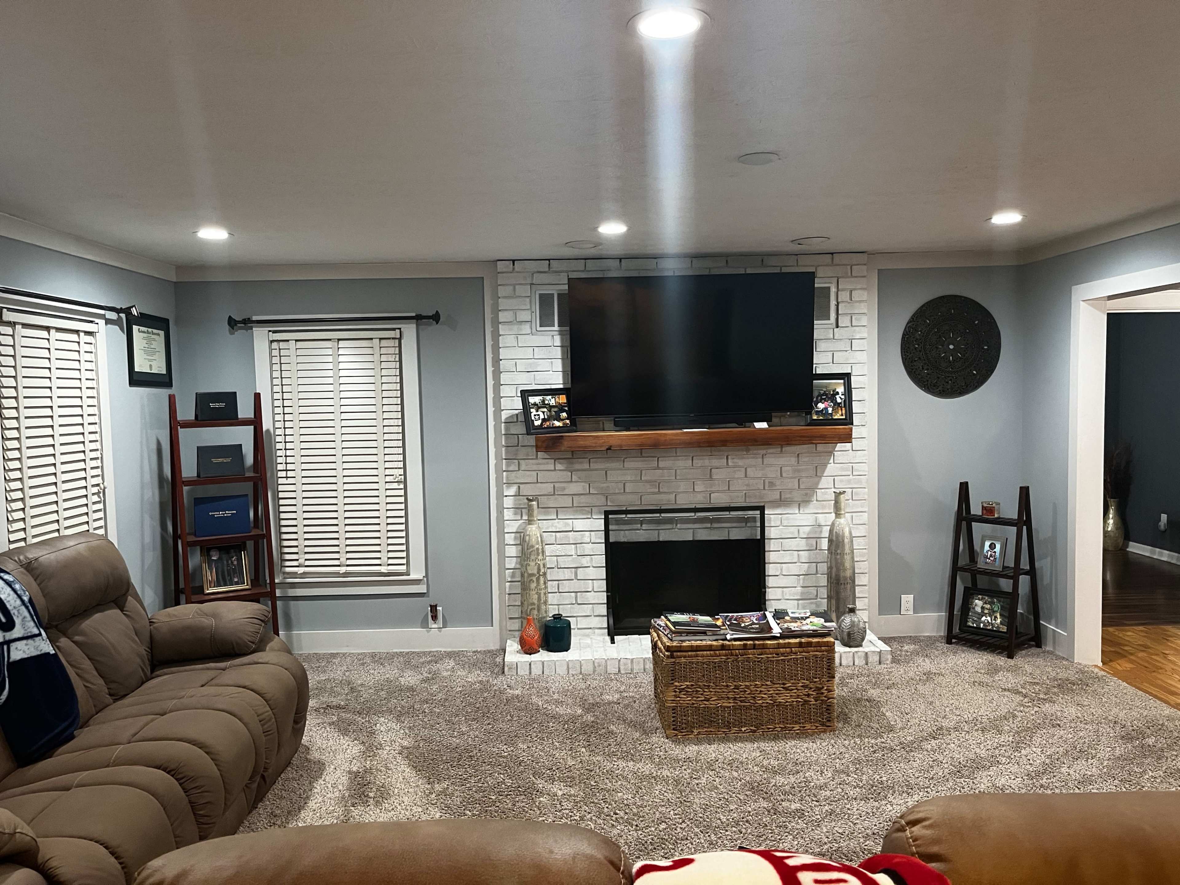 A living room with a light gray color scheme, featuring a white brick fireplace and a mounted television above a wooden shelf.