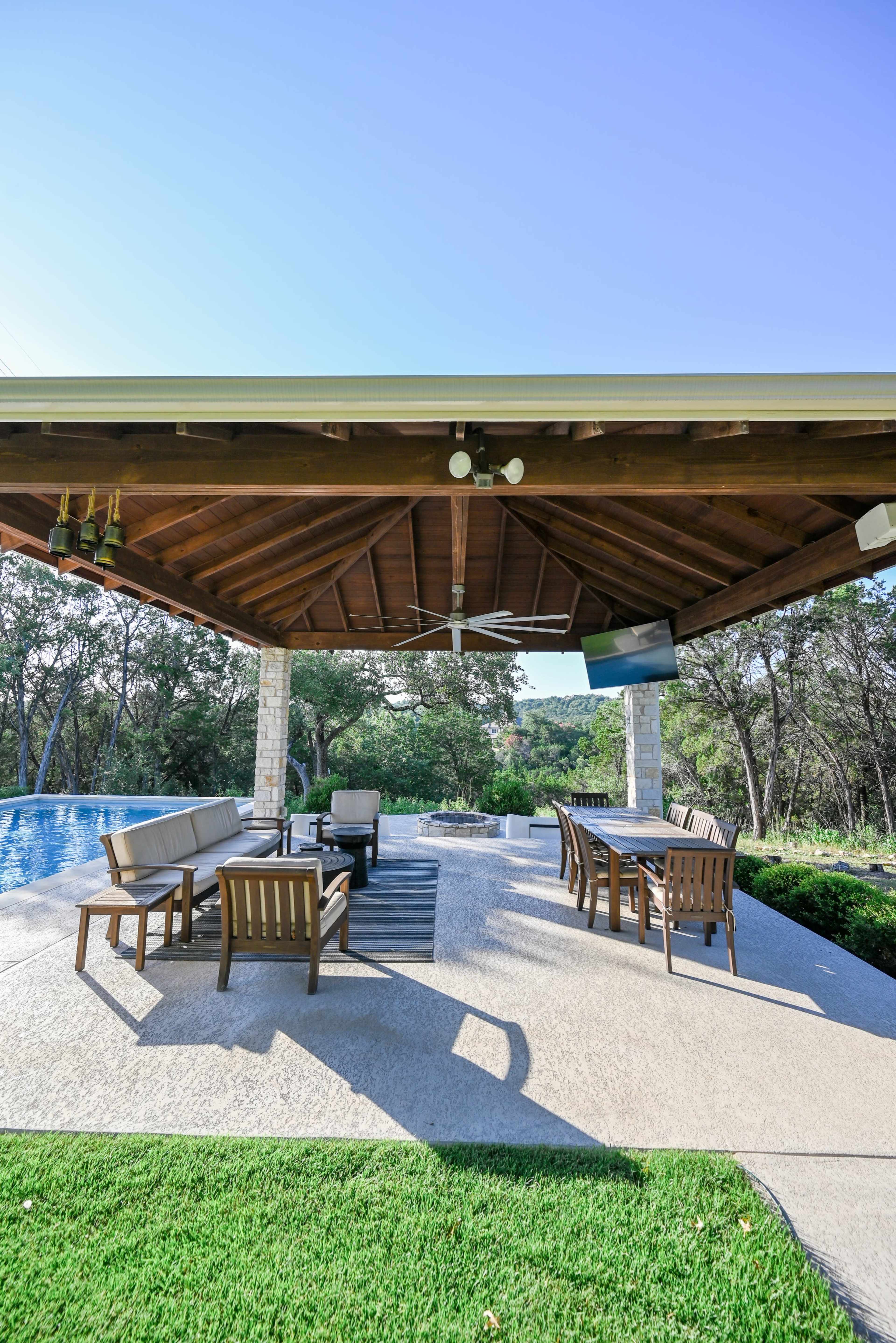 The image shows a covered outdoor patio with wooden furniture, a pool in the background, and a scenic view of trees.