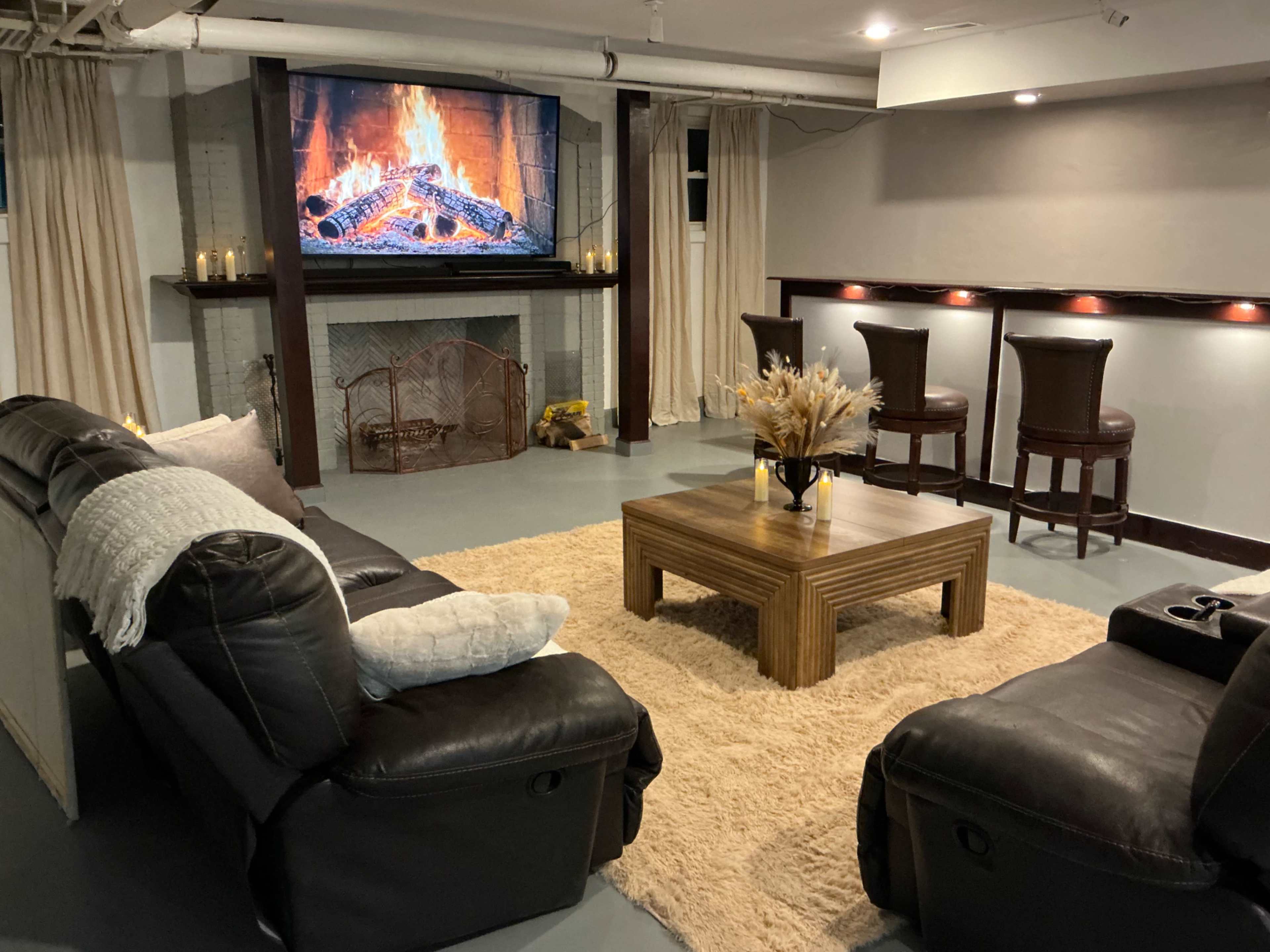 A cozy living room features a large television displaying a fireplace, a brown sofa set, a wooden coffee table, and bar stools along the back wall.