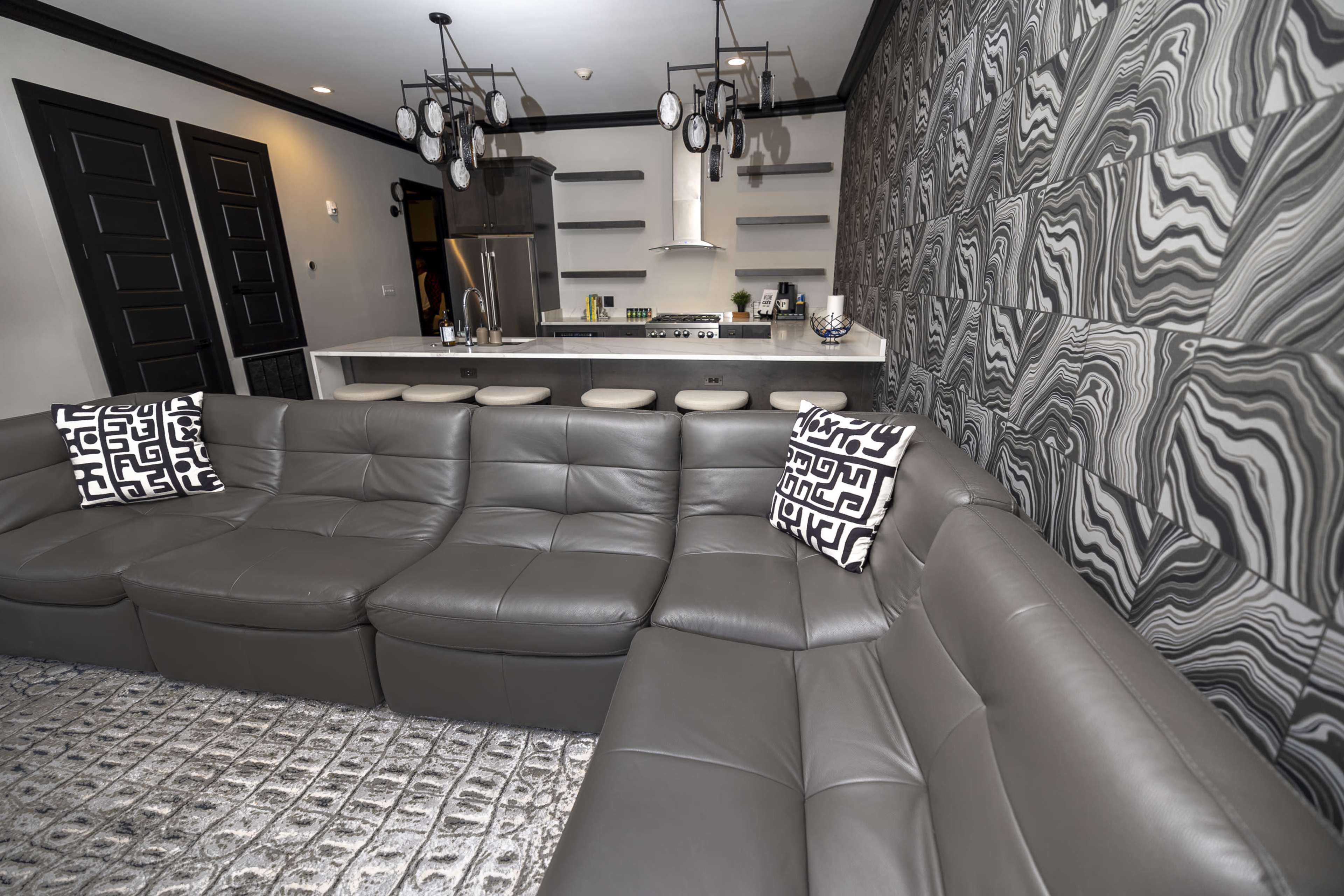 A modern living room features a gray sectional sofa, a patterned area rug, and a kitchen counter with sleek cabinetry and appliances in the background.
