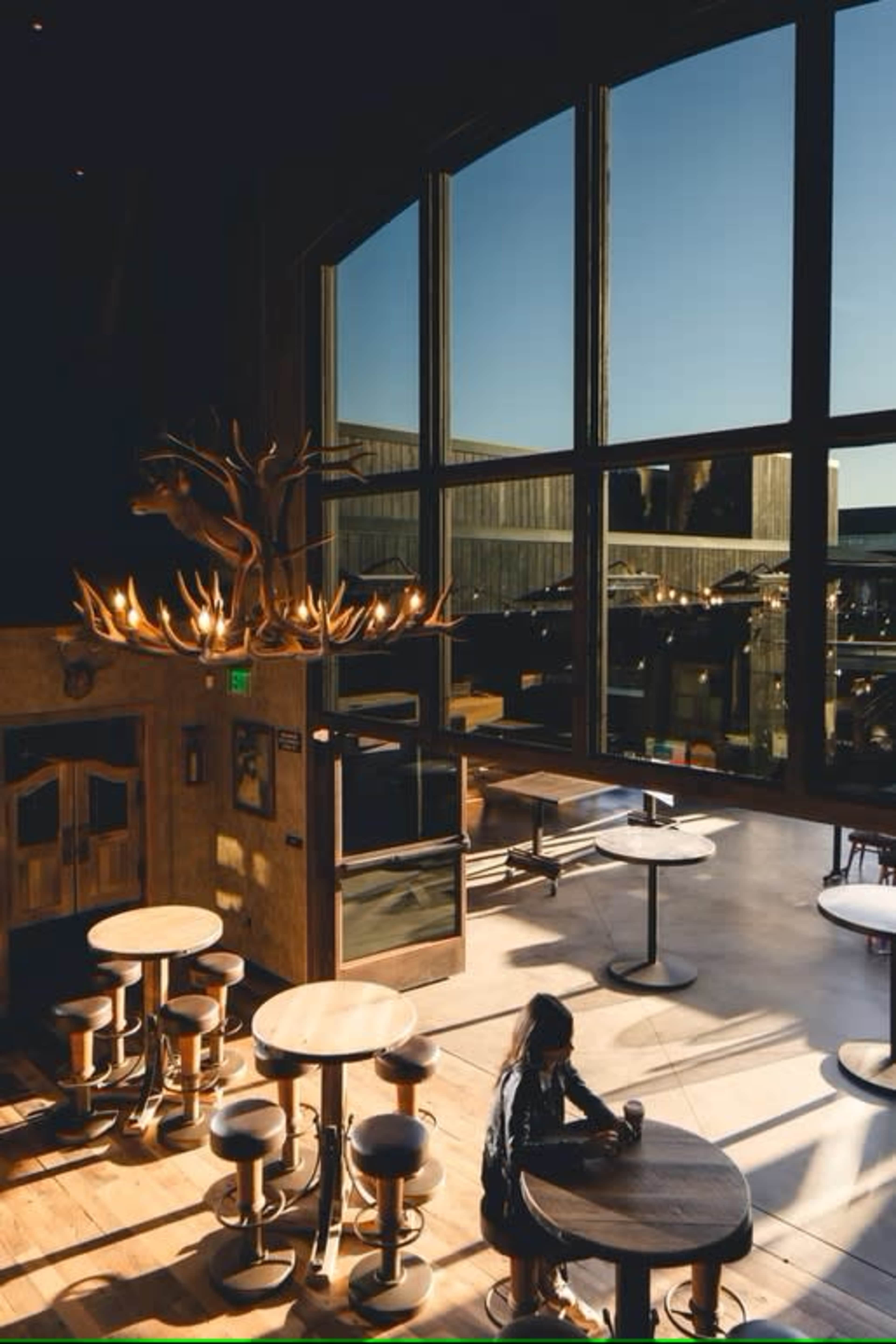 A well-lit café features large windows, wooden tables, and a decorative antler chandelier.