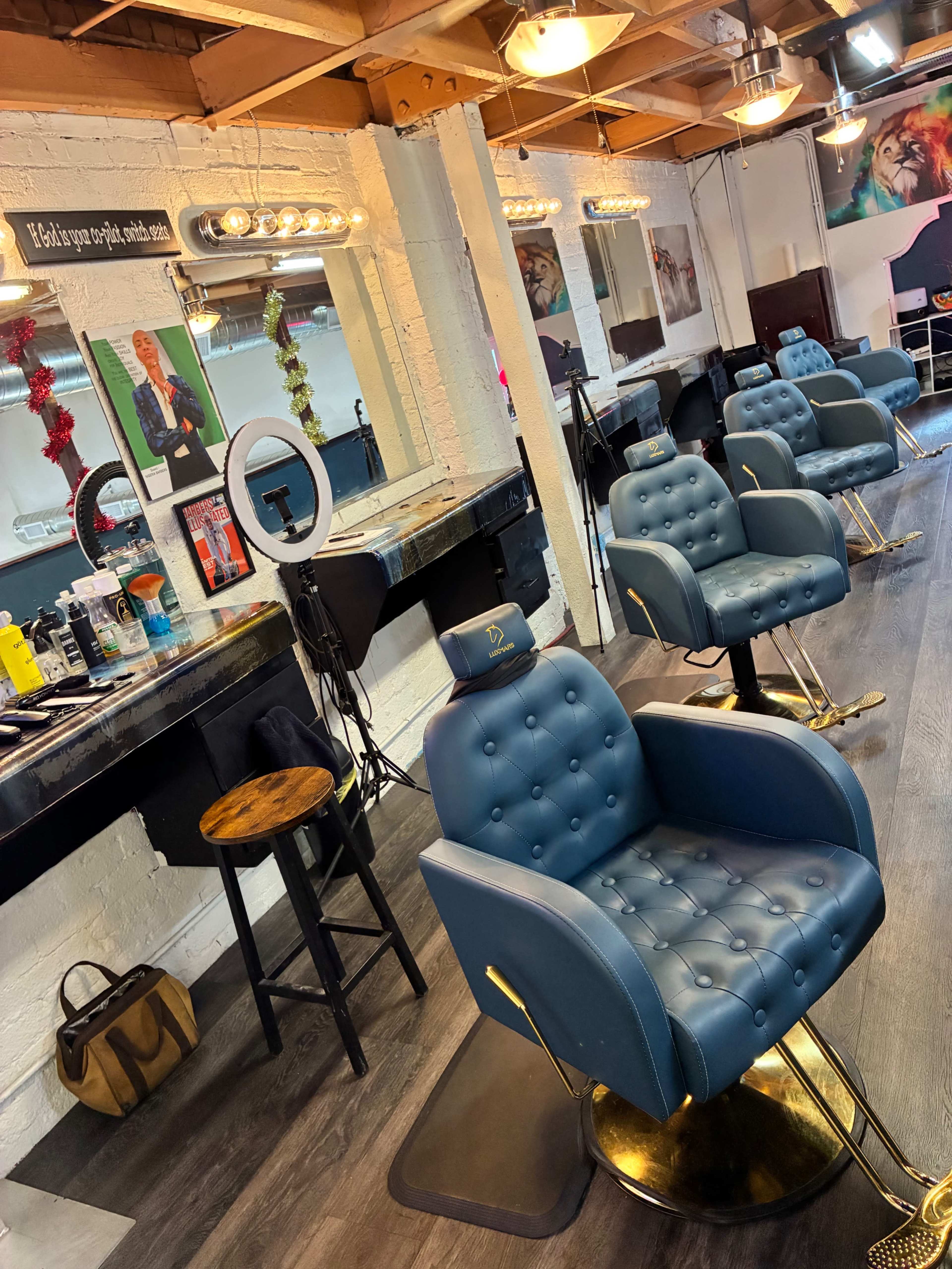 The image shows a modern barbershop interior with several blue, cushioned barber chairs and well-lit mirrors arranged along a brick wall.