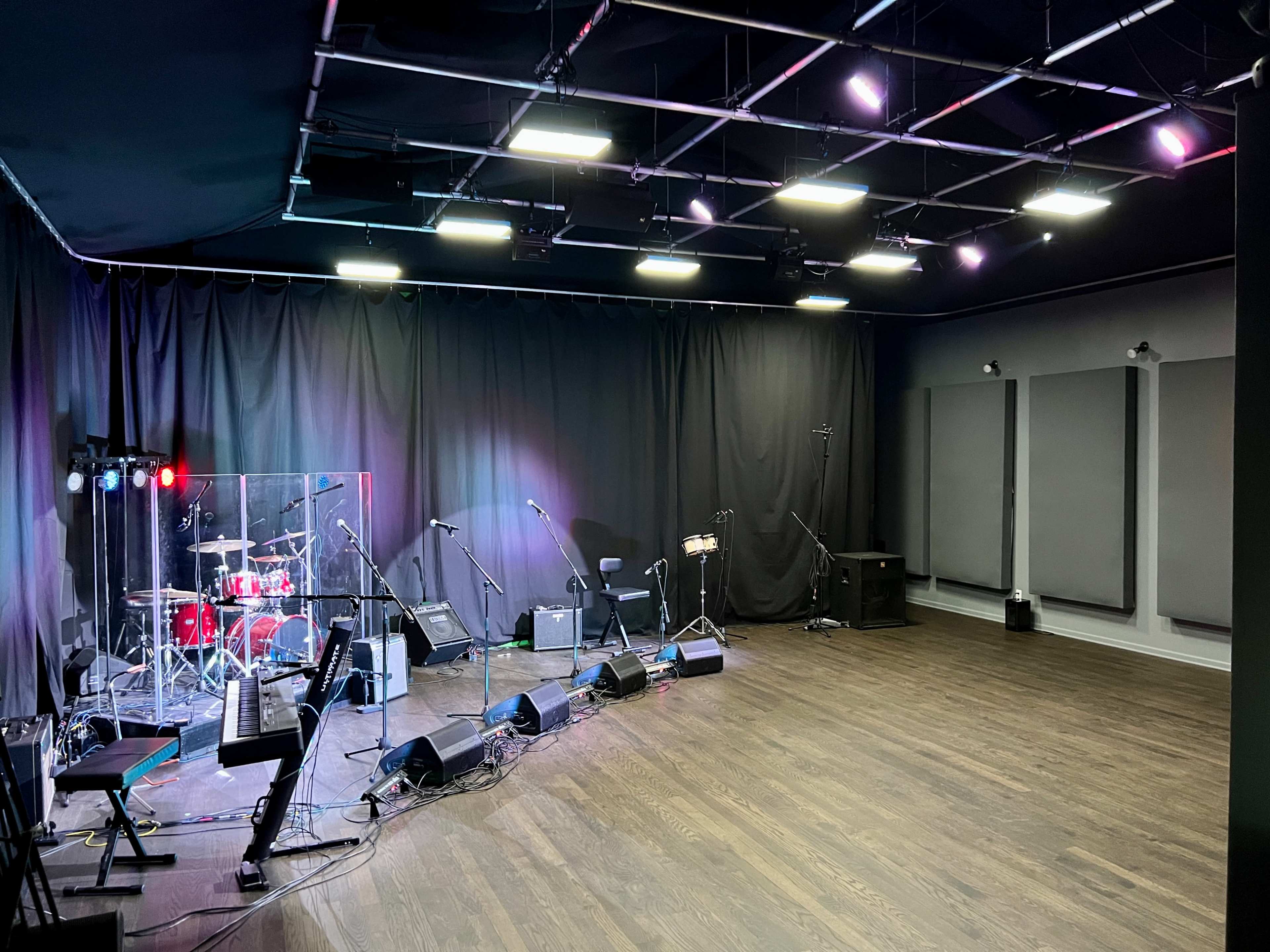 An empty music rehearsal space featuring a drum set, microphones, amplifiers, and sound equipment set against black curtains and acoustic panels.
