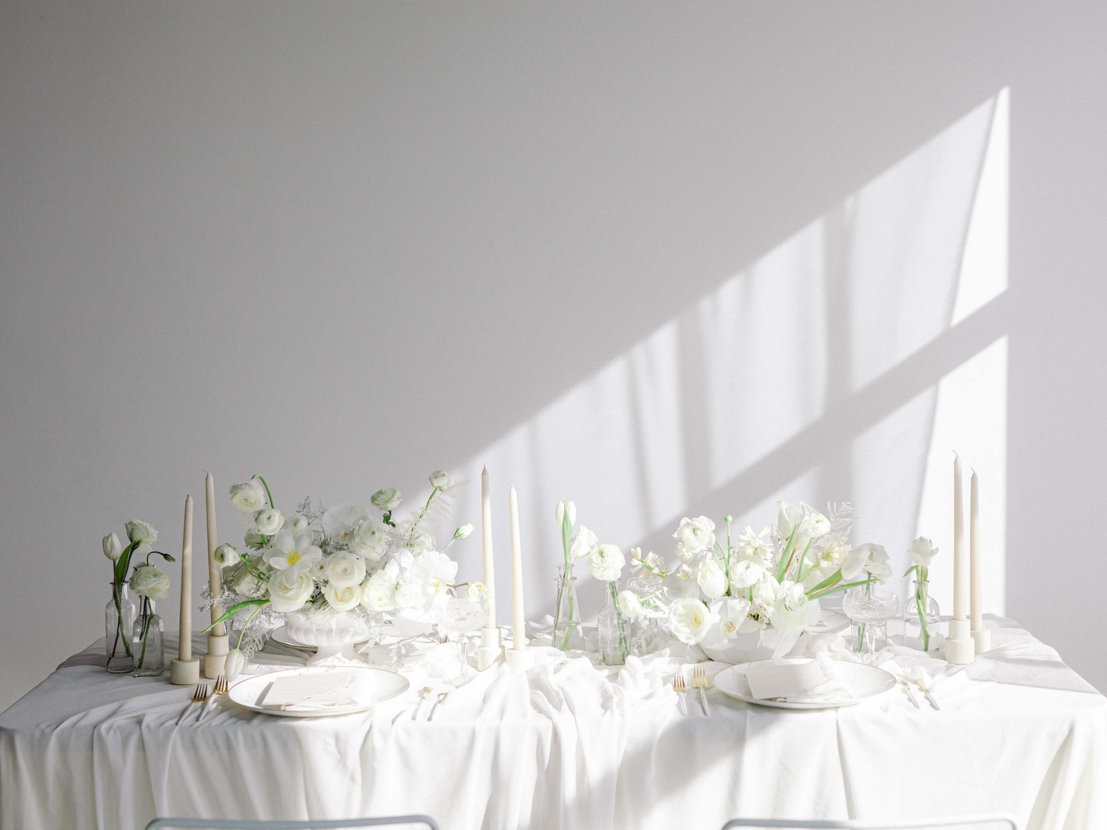 A dining table is set with white tablecloths, floral centerpieces, candle holders, and plates under soft, natural light.