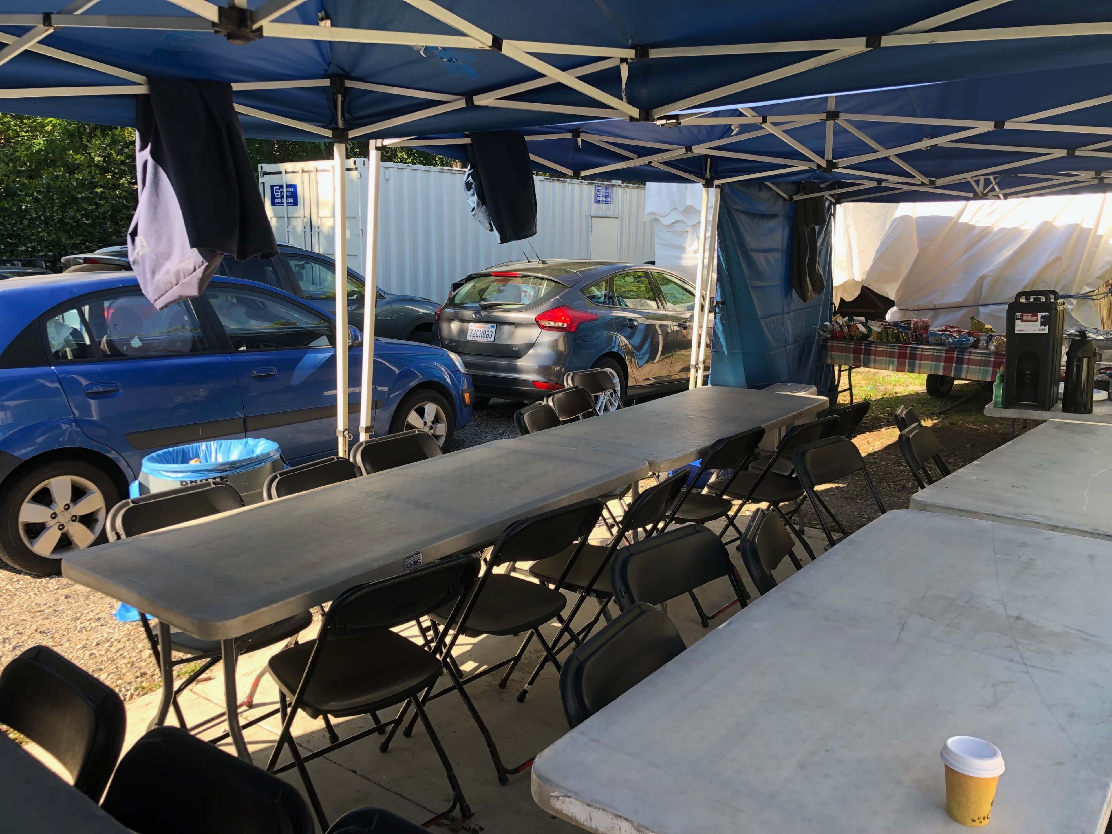 A row of long tables and chairs is set up beneath blue tents, with parked cars visible in the background.