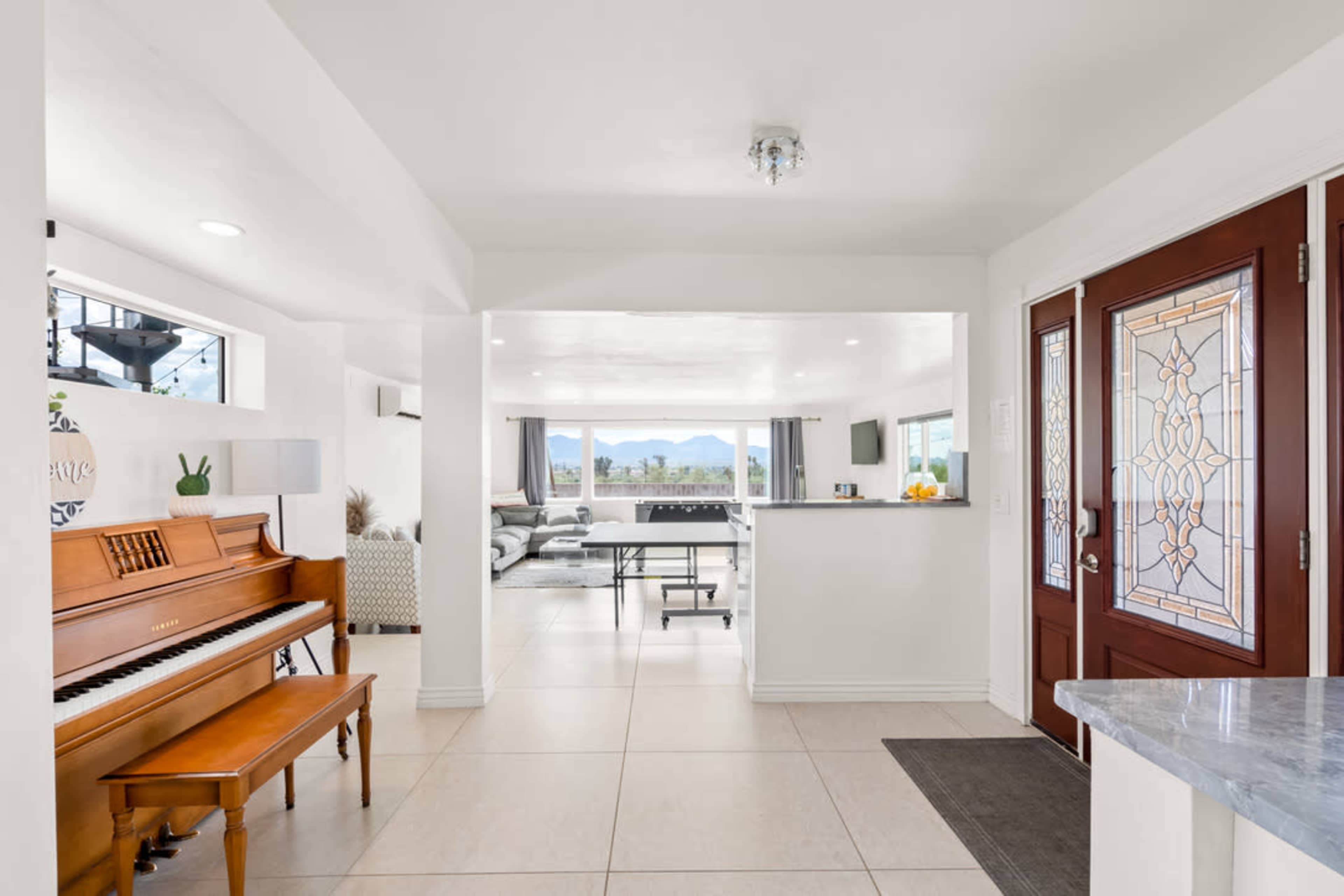 A bright entrance area featuring a wooden piano, an open floor plan leading to a living space with large windows, and a door with decorative glass.