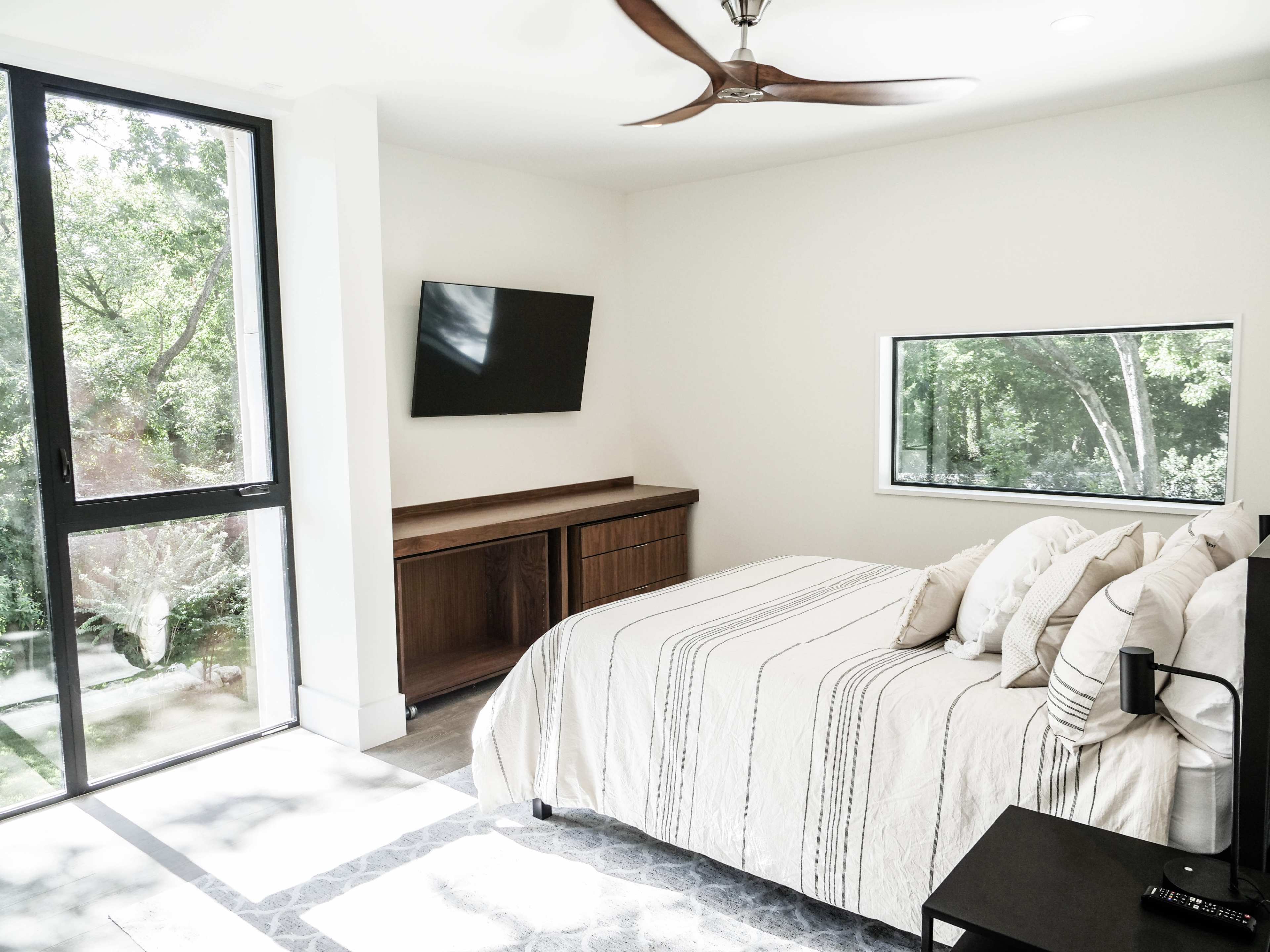 A modern bedroom with a bed, a wall-mounted television, and a window overlooking a lush outdoor area.