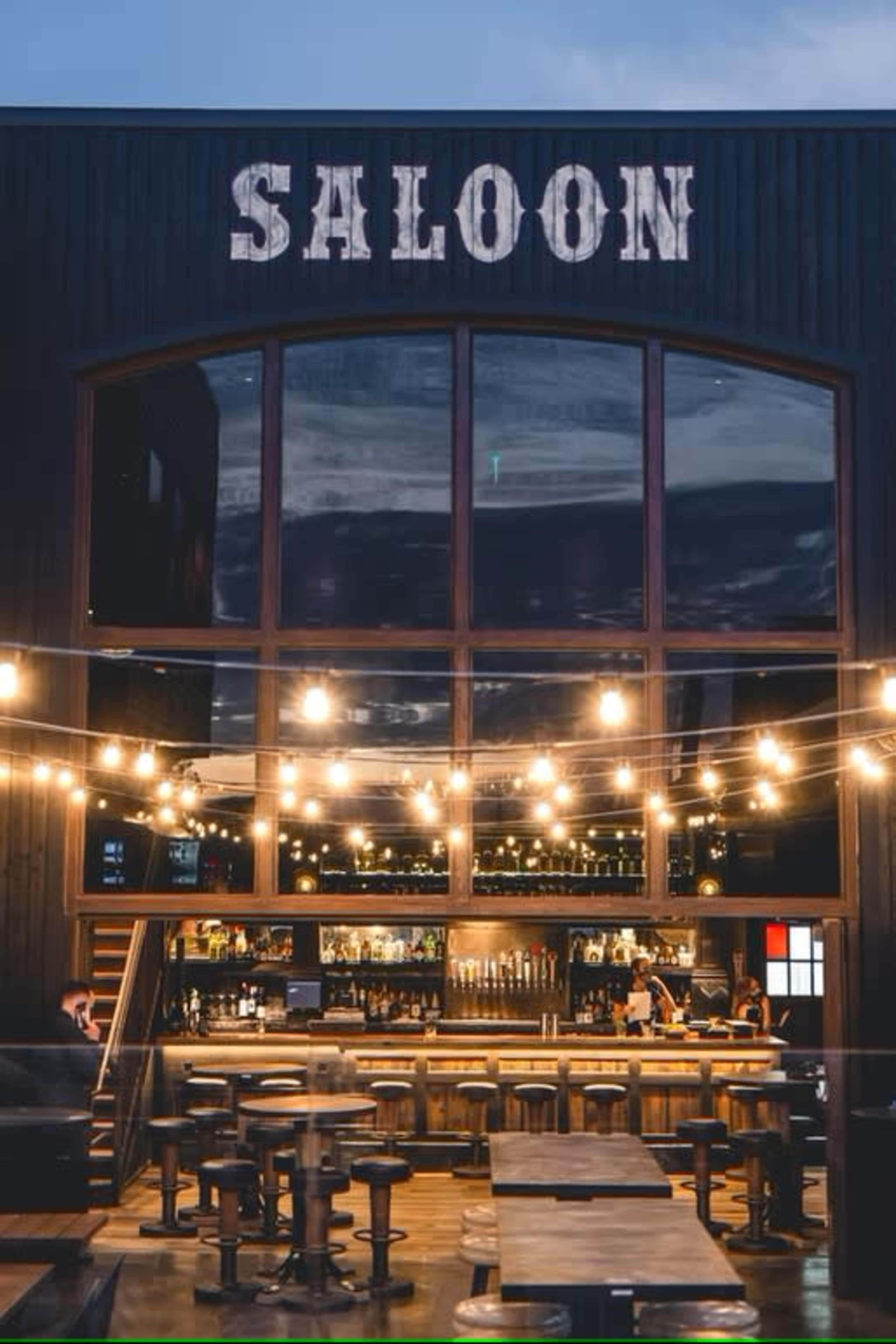 The image shows a rustic saloon with large windows, a bar area illuminated by string lights, and wooden seating arrangements.