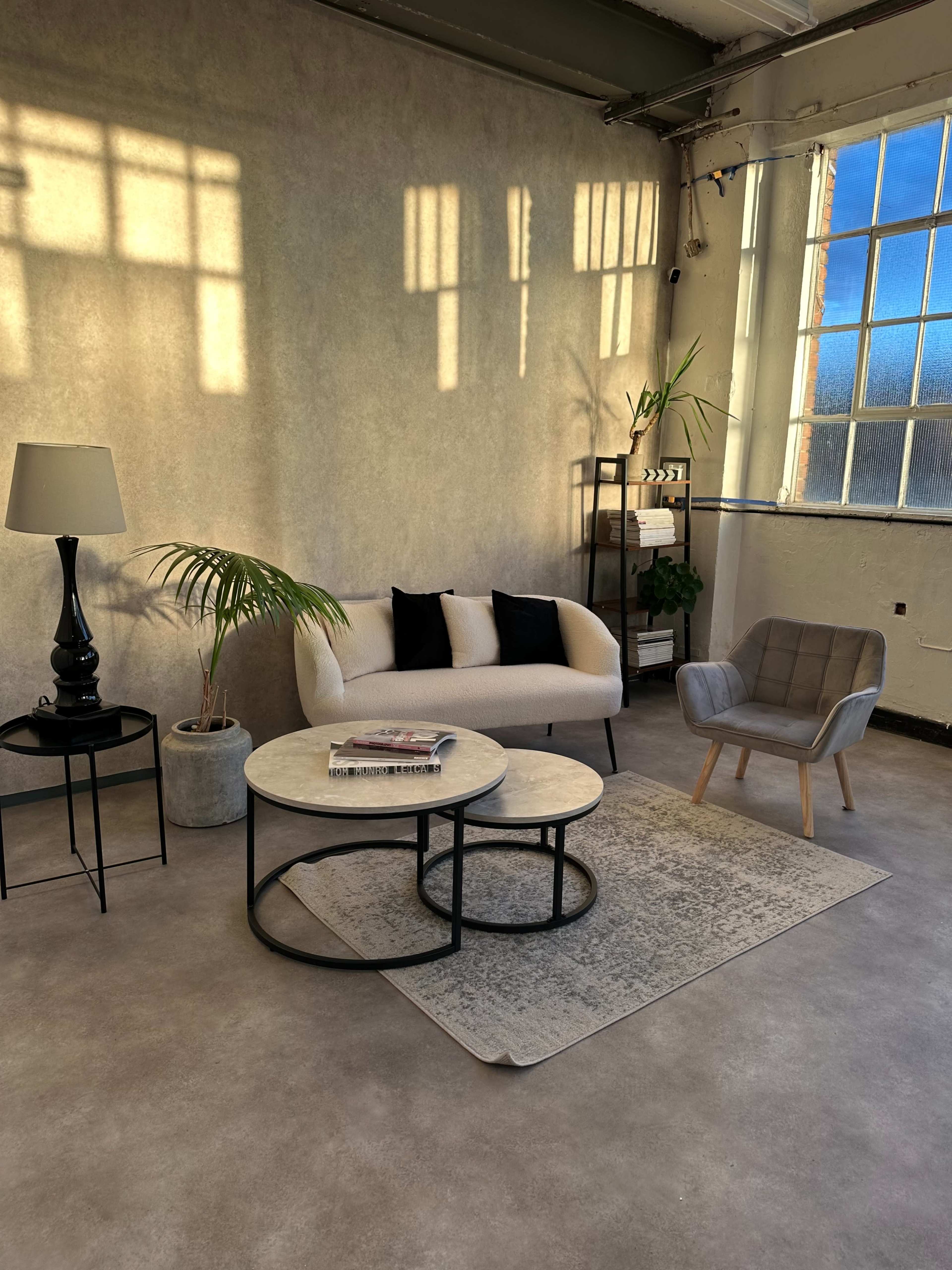 A cozy living area features a beige sofa with black pillows, two round coffee tables, a floor lamp, and a modern armchair, all set against a textured wall and large window.