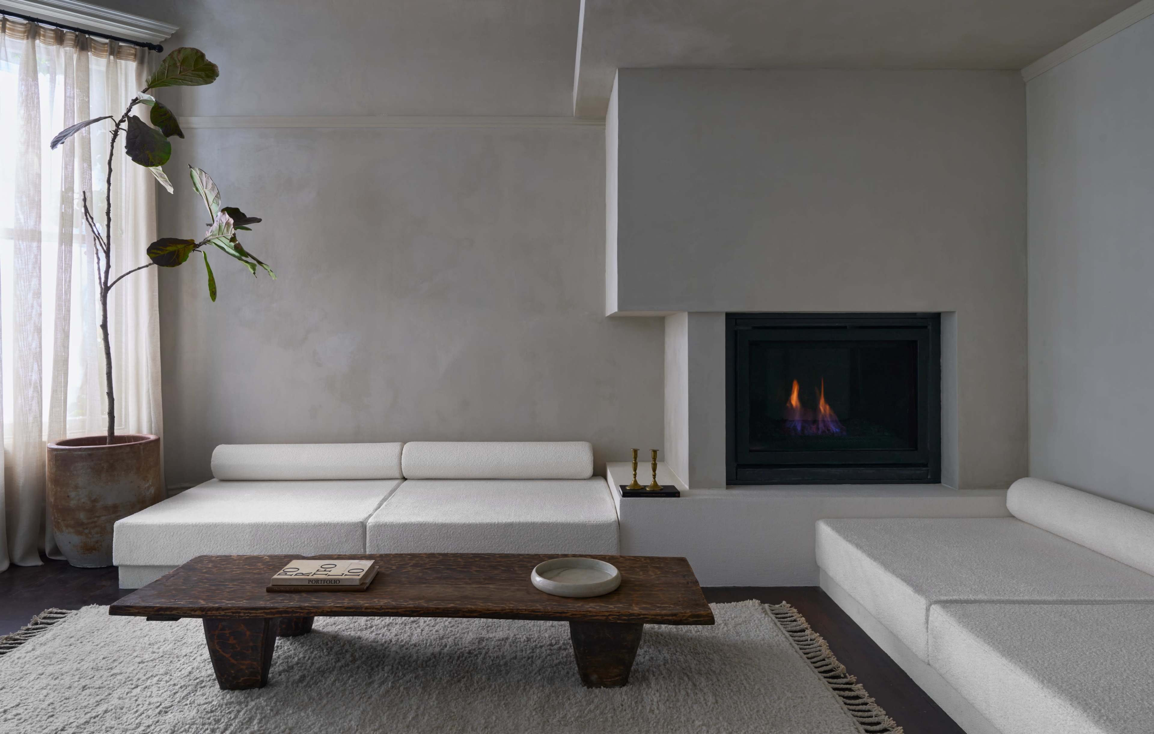 A minimalist living room features a white sectional sofa, a rustic wooden coffee table, and a modern fireplace with a small flame.