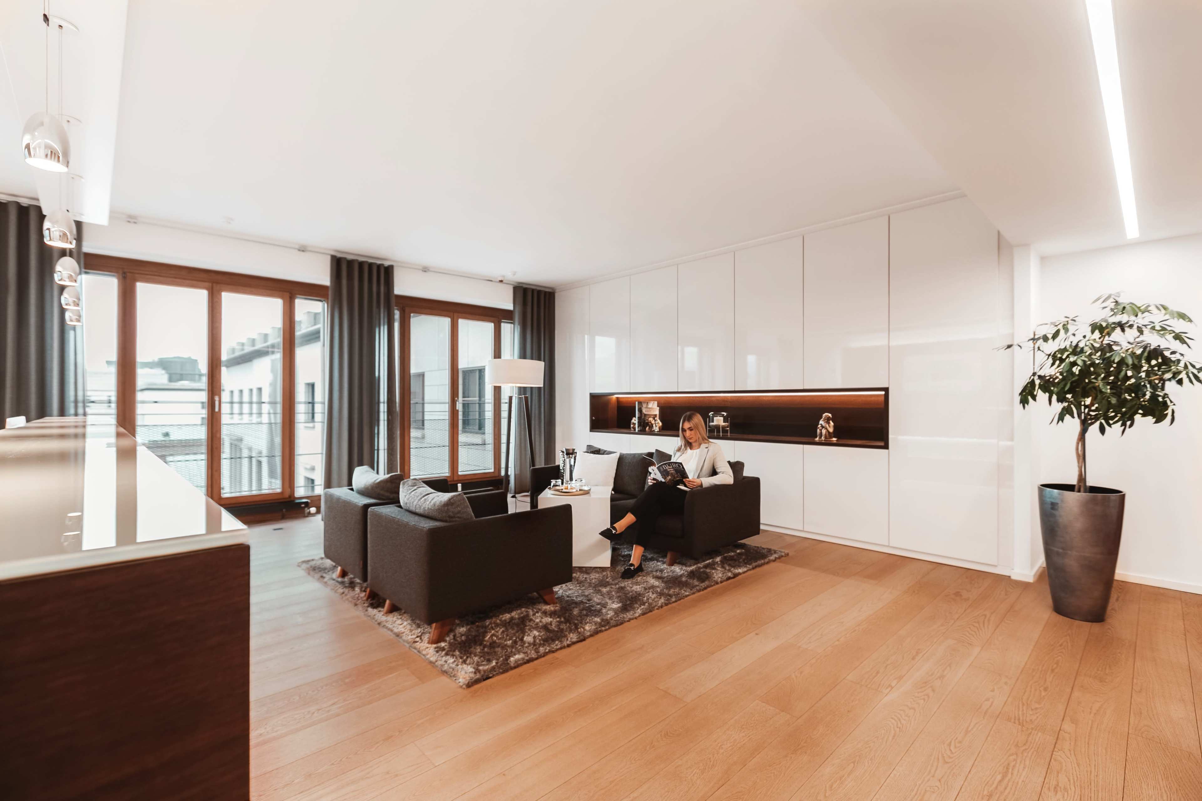 A woman sits on a couch in a modern living room with large windows and minimalist décor.