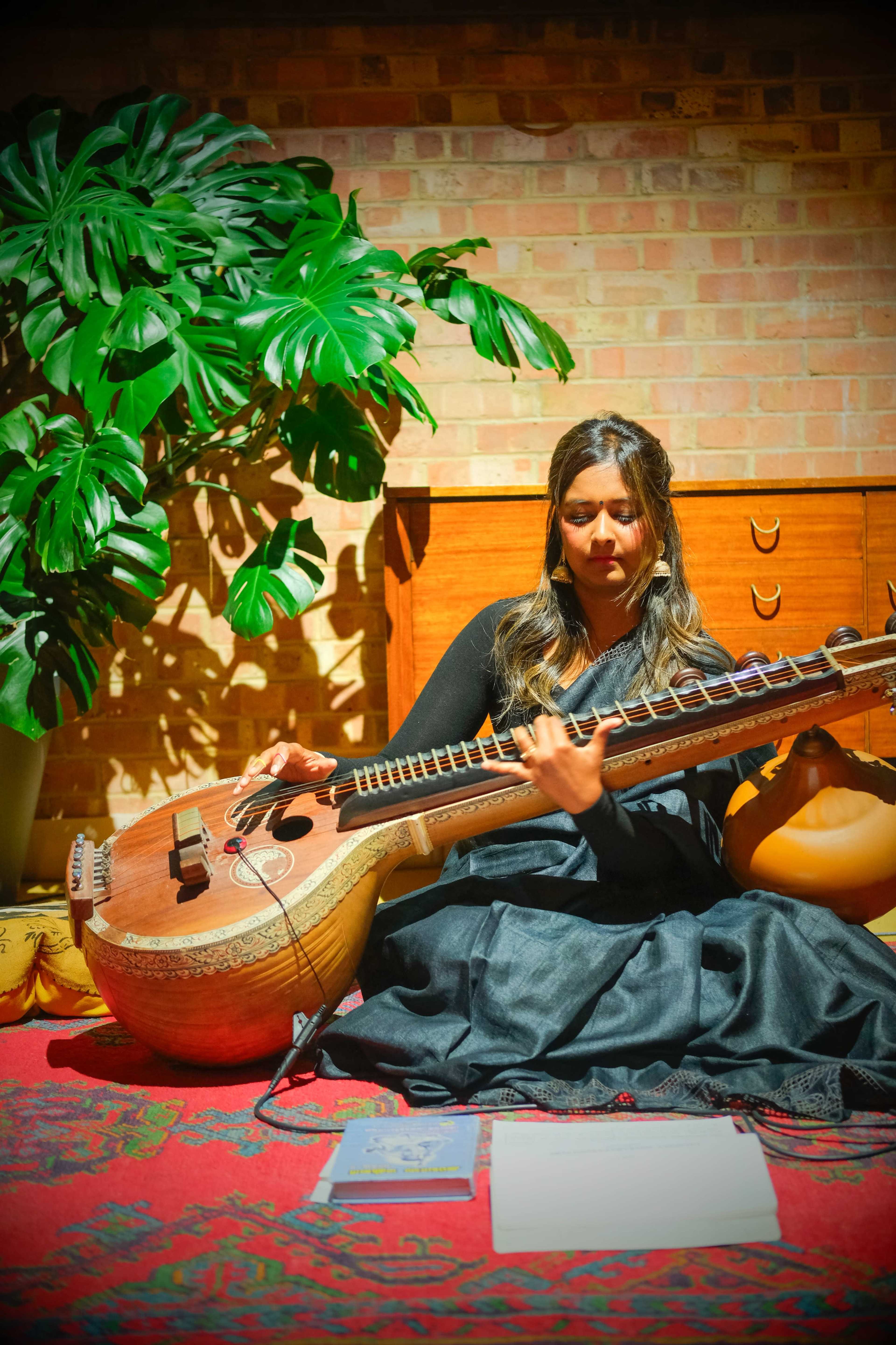 A woman playing a stringed instrument sits on a patterned rug in front of a leafy plant and wooden furniture.