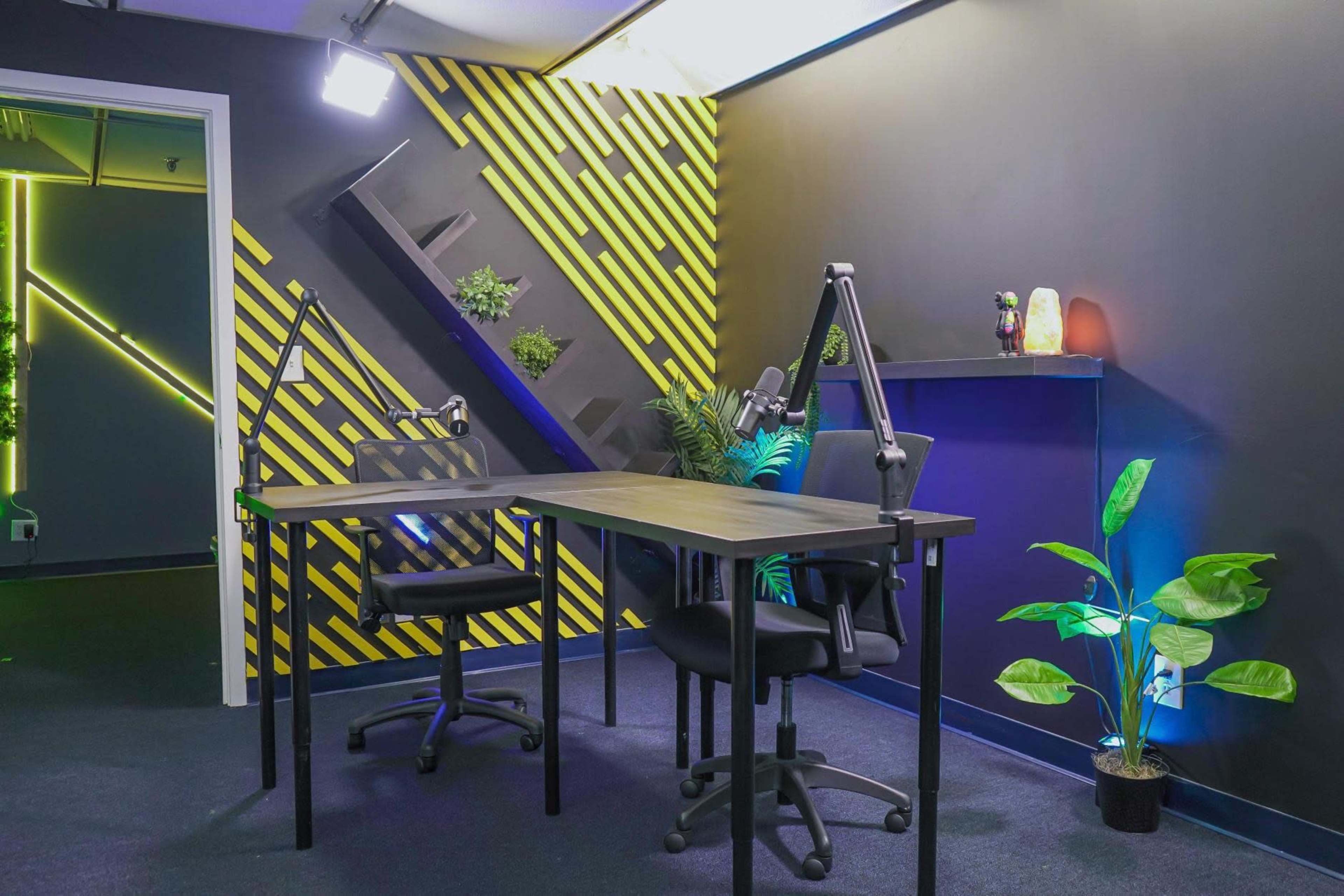 The image shows a modern workspace with two black chairs and a table, set against a backdrop of yellow and black diagonal stripes and greenery.