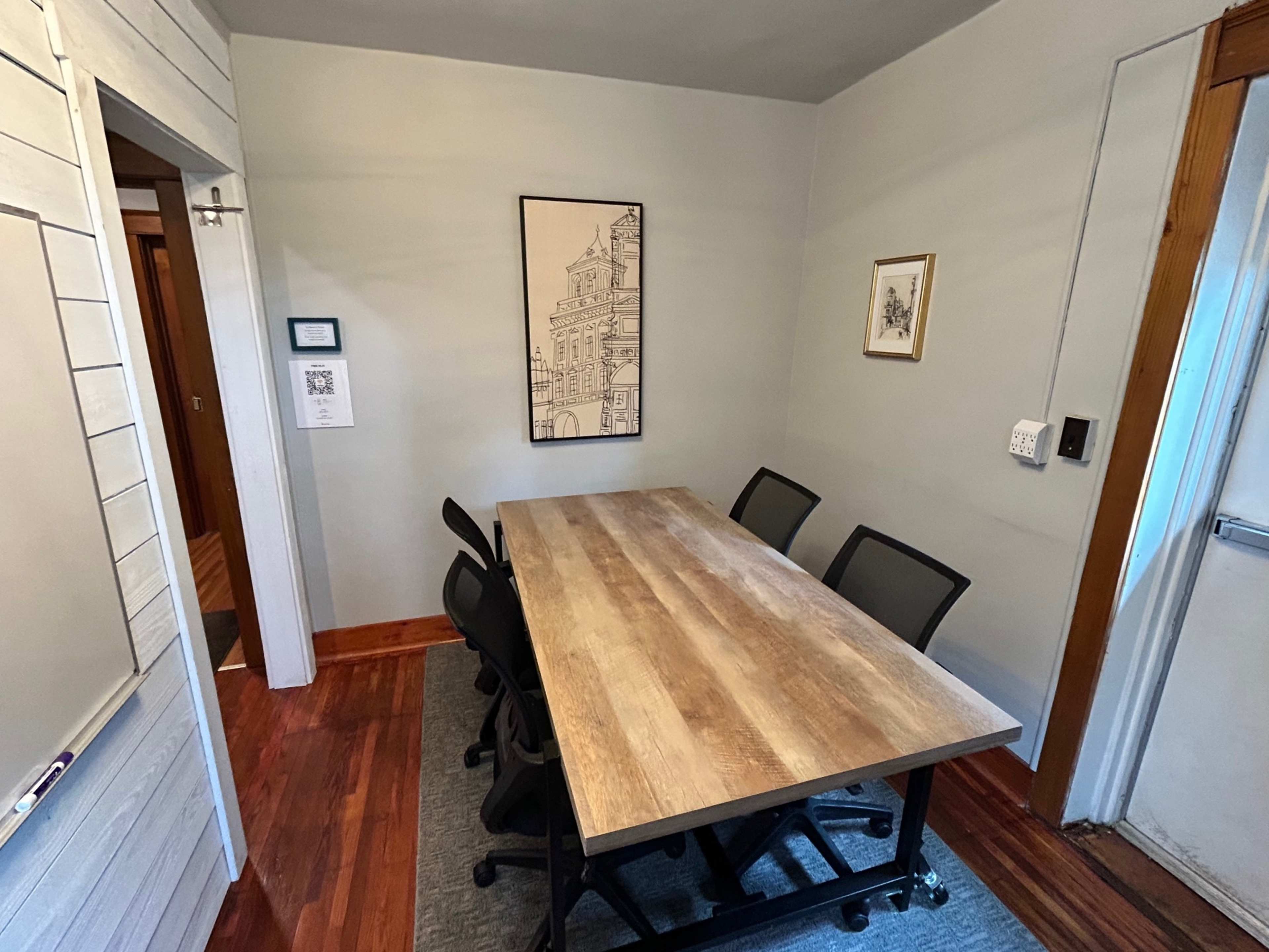 The image shows a small meeting room with a wooden table surrounded by black office chairs, a wall-mounted QR code sign, and framed artwork on the walls.