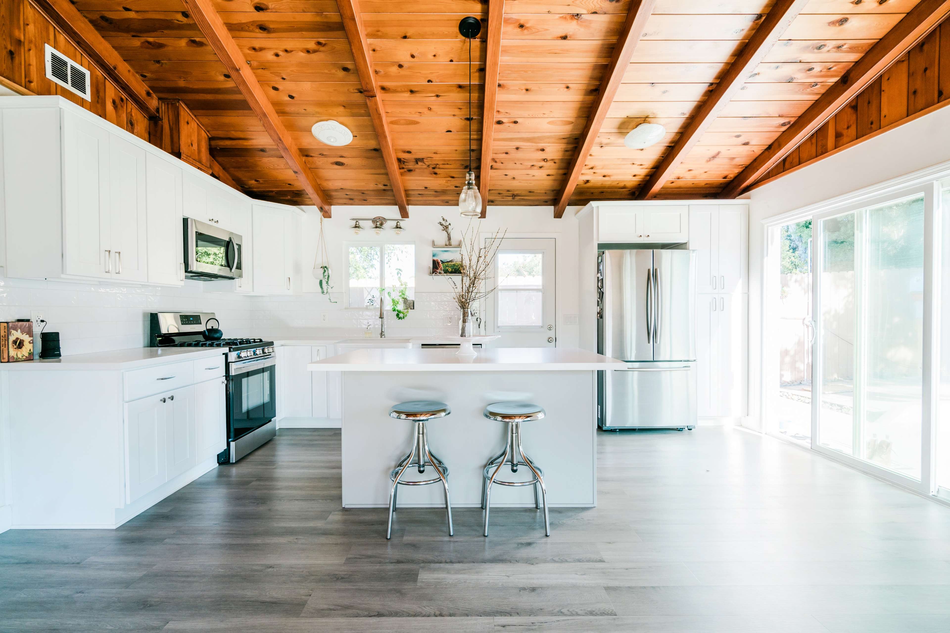 The image shows a modern kitchen with white cabinetry, a central island with two metal stools, and exposed wooden beams on the ceiling.