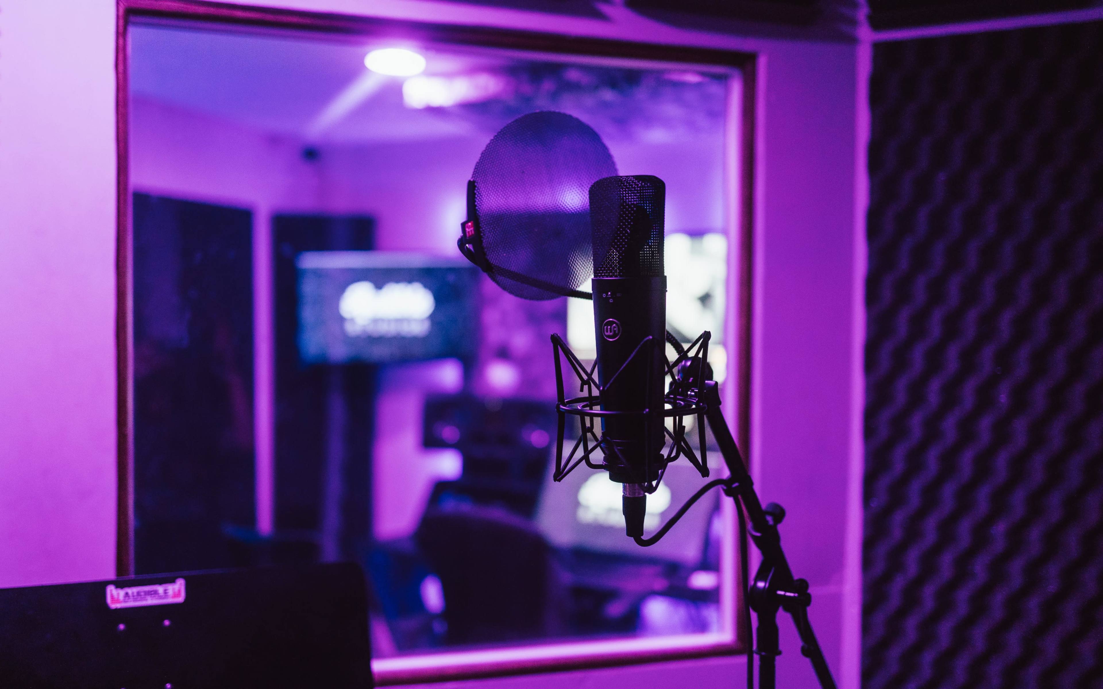 A microphone stands in front of a soundproof booth, with a purple hue illuminating the scene.