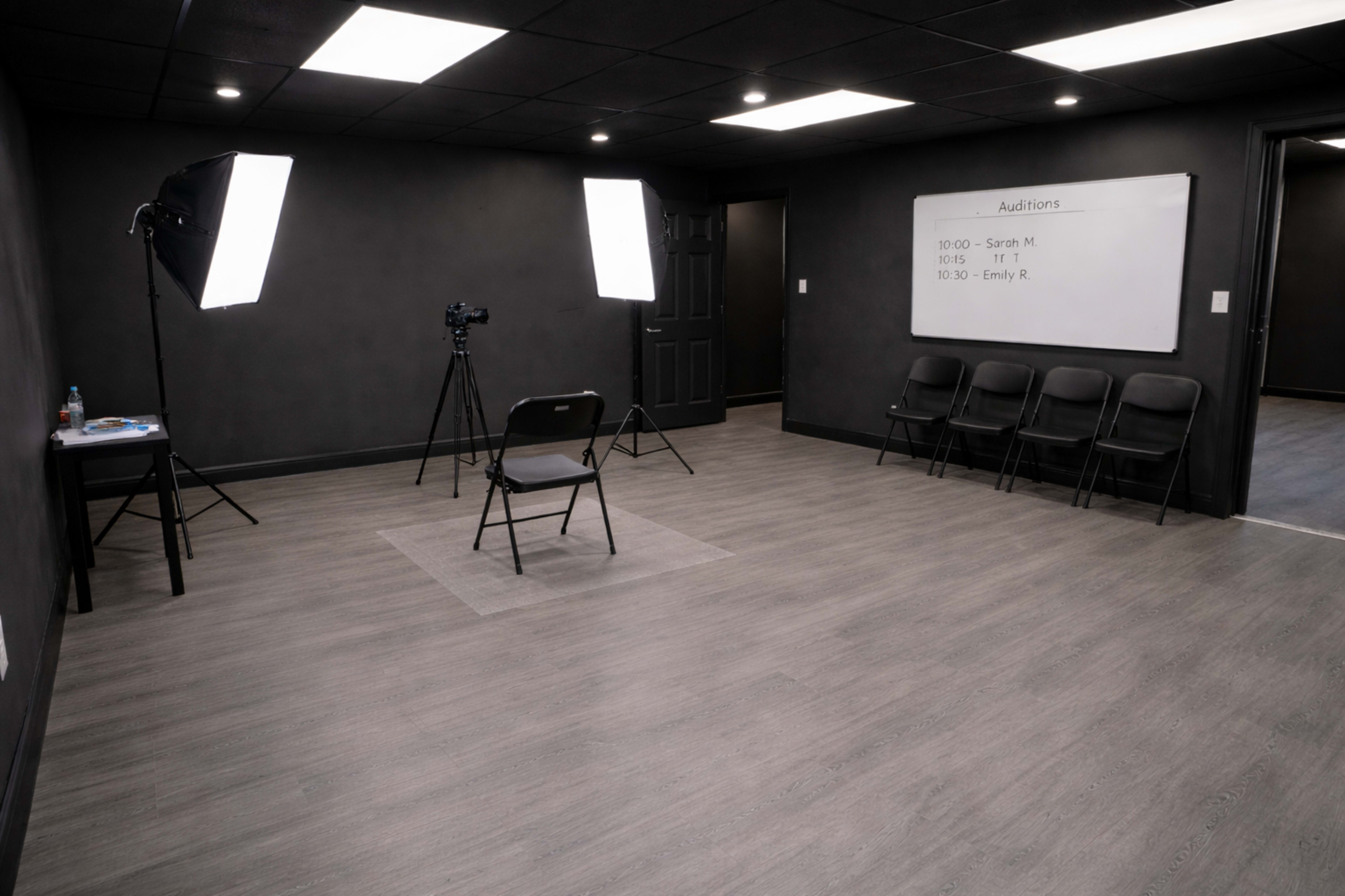 The image shows a dimly lit audition room with a single chair in front of a camera and bright lights, along with a schedule on a whiteboard listing audition times.