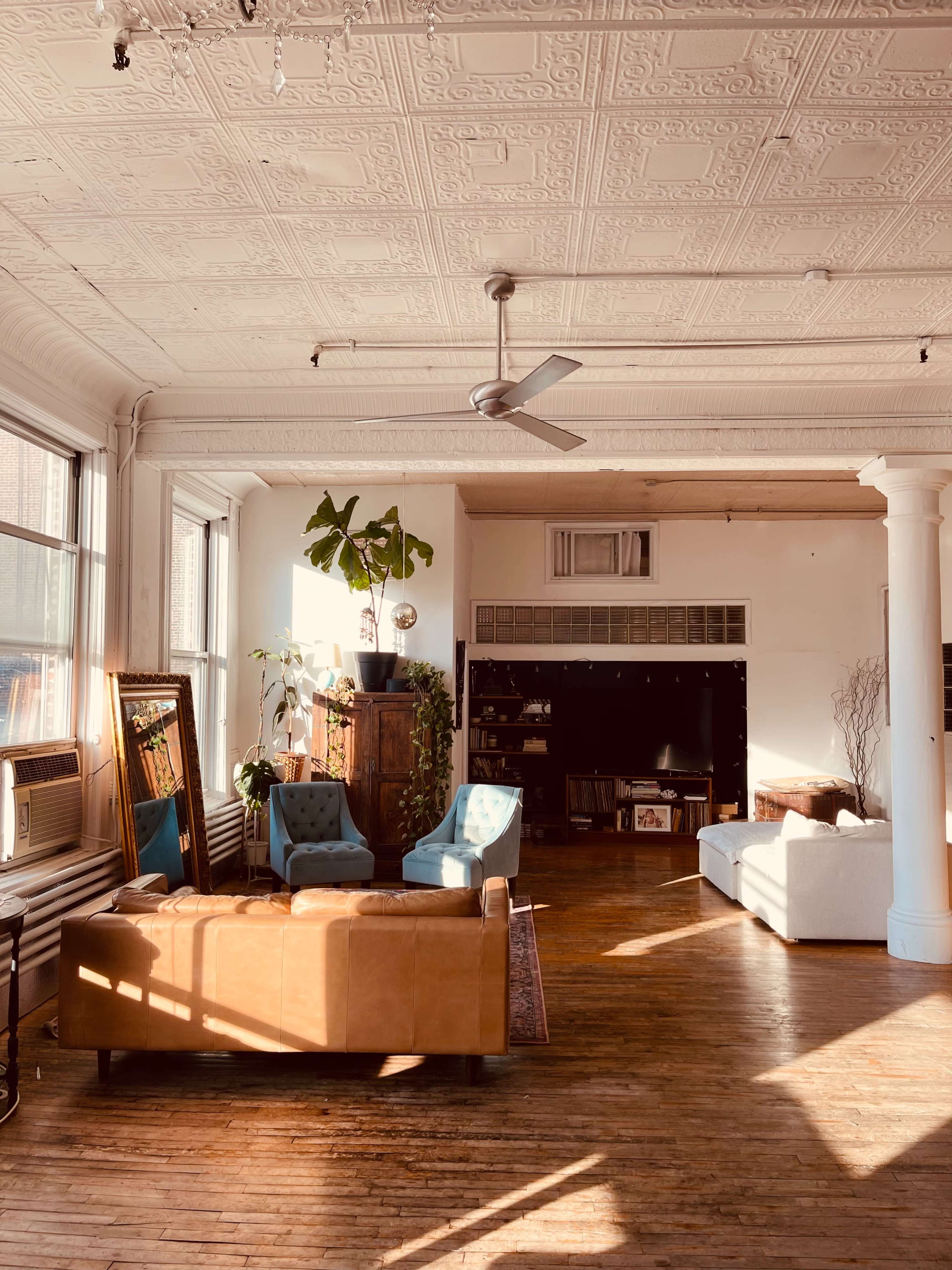 The image shows a well-lit, spacious living area featuring a mix of furniture, including a leather couch, blue chairs, and a white sofa, along with a large plant and decorative accents in a room with high ceilings and ornate details.