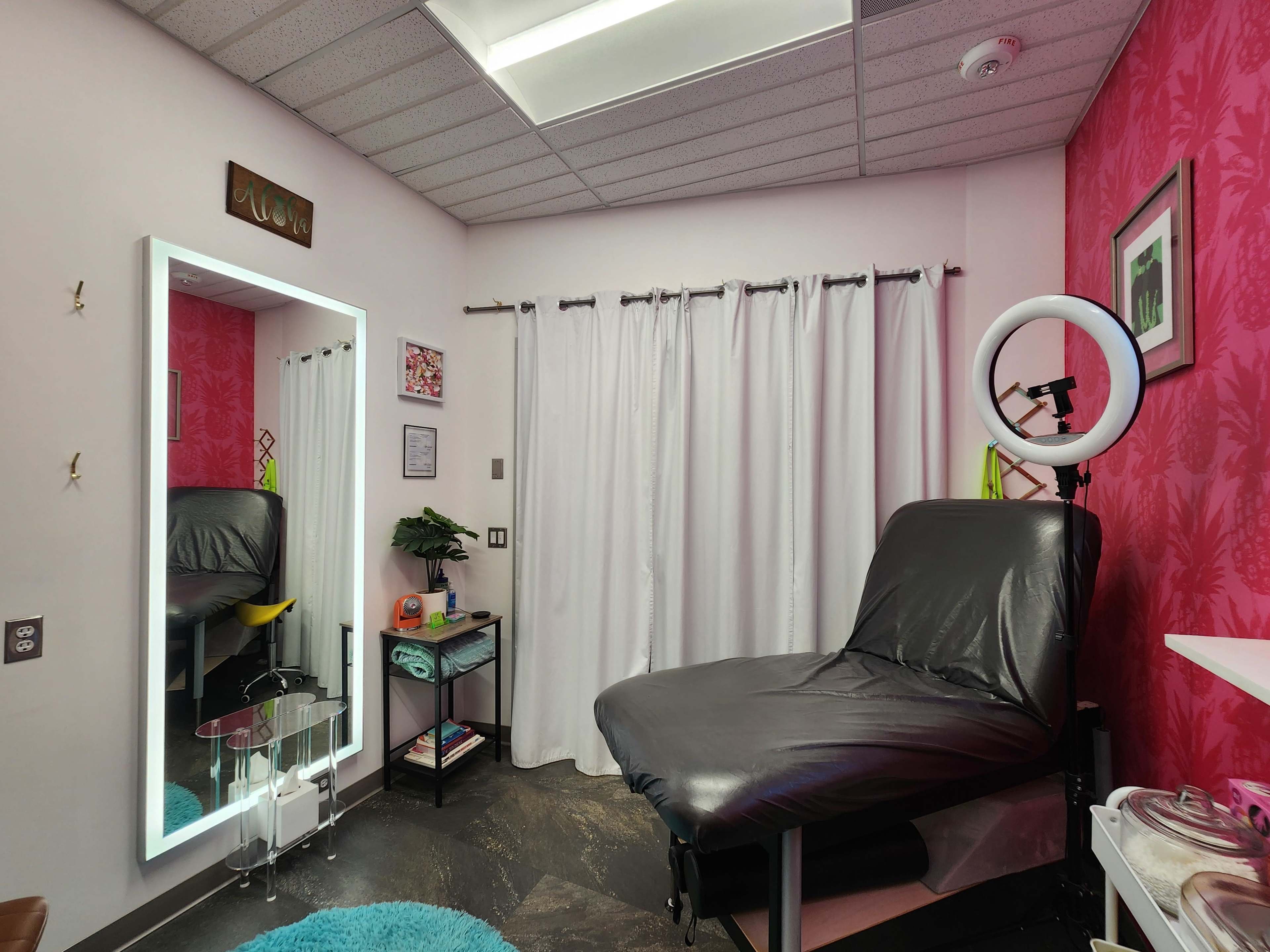 The image shows a brightly colored treatment room with a black examination bed, a circular mirror light, and white curtains.