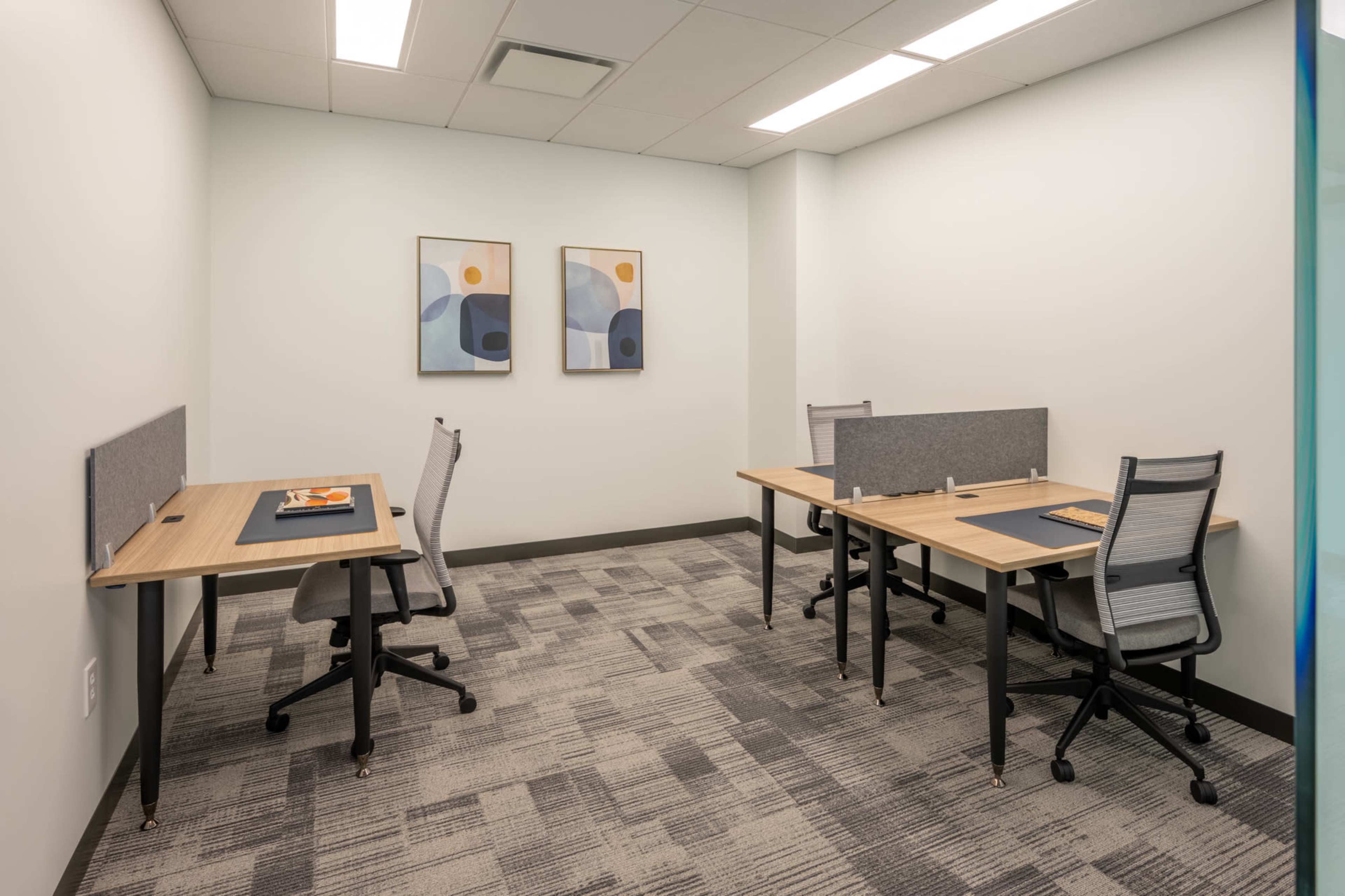 The image shows a small office space with two wooden desks, each equipped with a chair and partition, and framed abstract art on the walls.