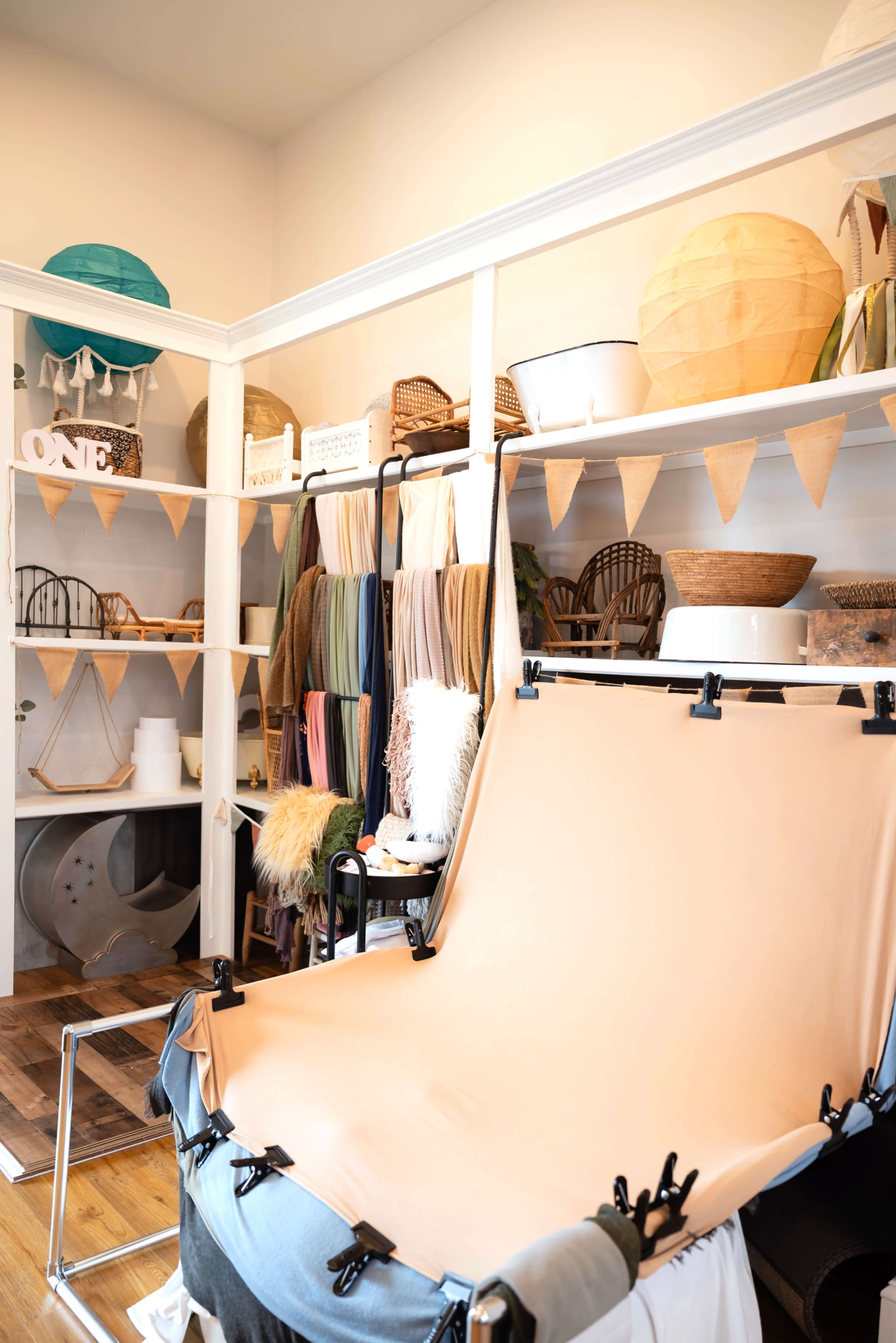 The image shows a well-organized studio space with shelves filled with various decorative items, fabrics, and craft supplies, and a backdrop setup in the foreground for photography or videography.