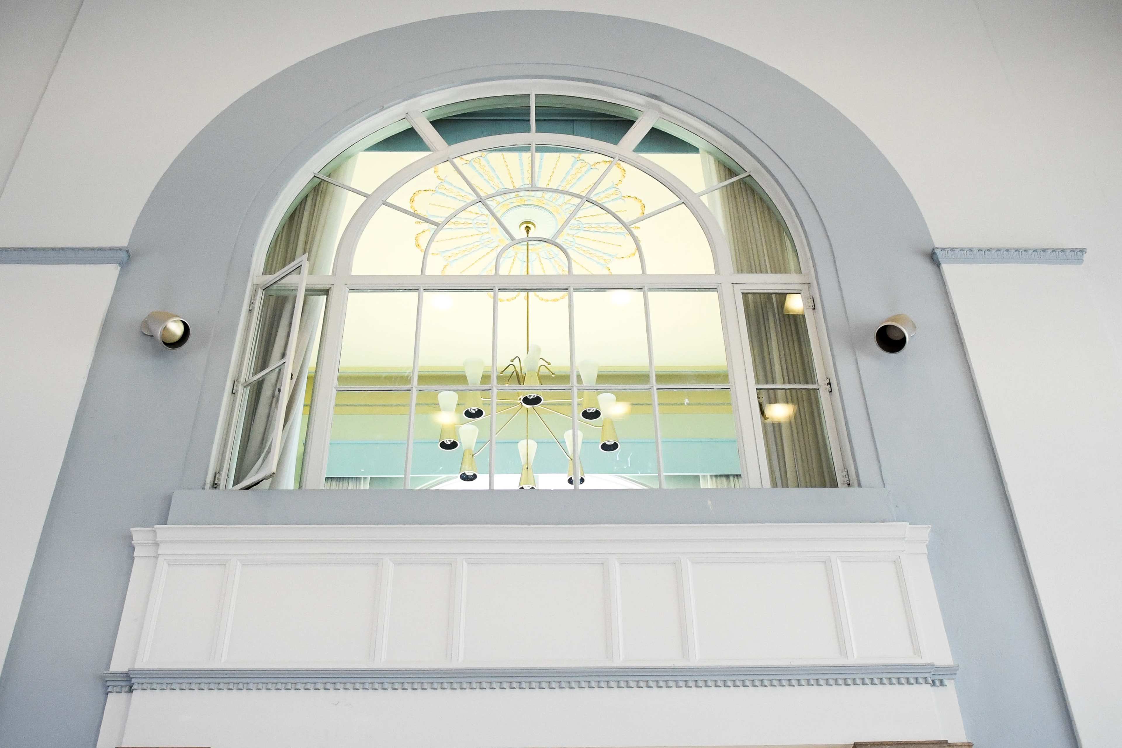 The image shows a large arched window with a chandelier visible through it, framed by a white wall and blue accents.
