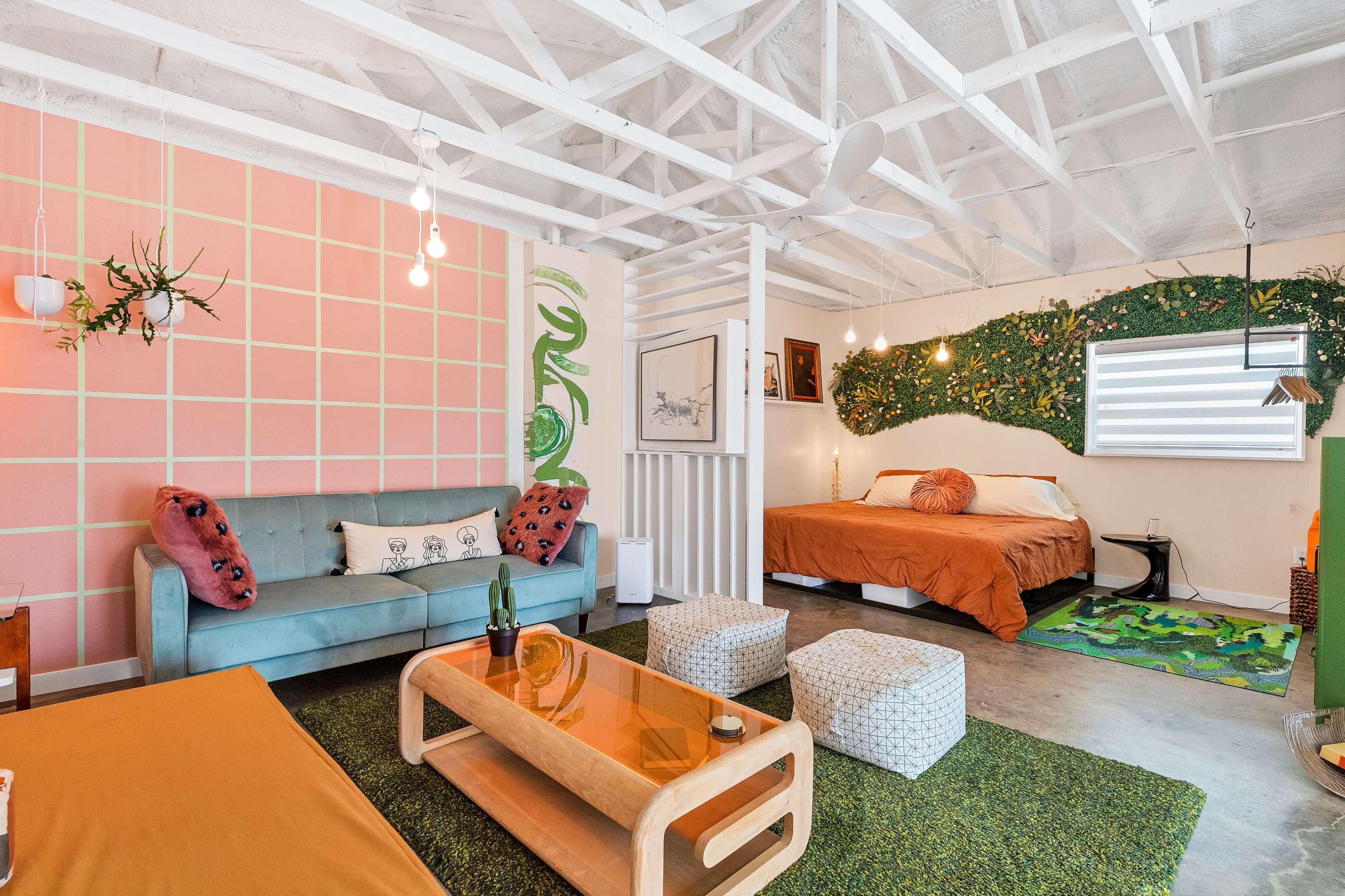 A brightly colored living space featuring a green sofa, a wooden coffee table, a bed with an orange duvet, and decorative plants on the walls.
