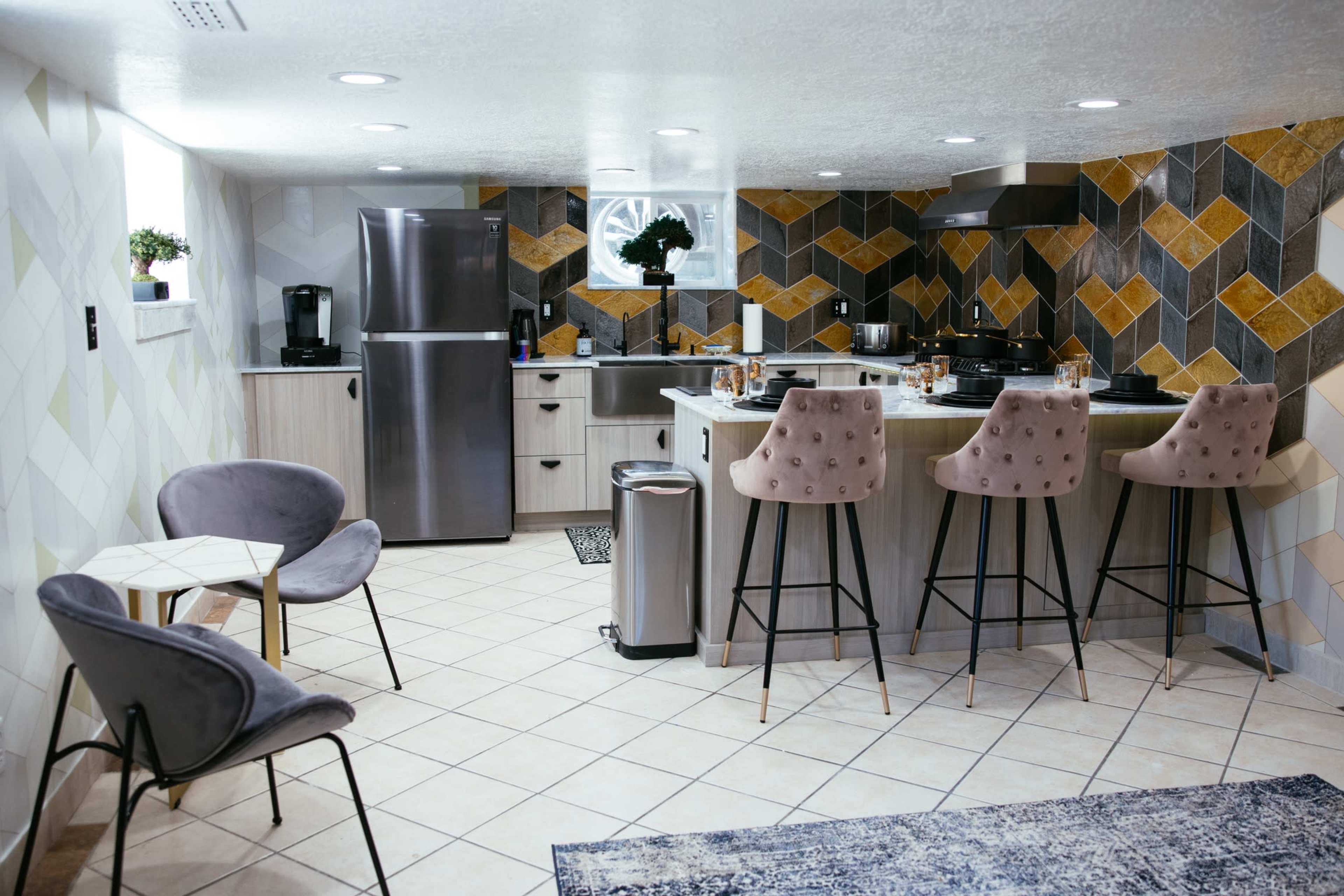 A modern kitchen with a patterned tile backsplash, stainless steel appliances, a dining area with bar stools, and a seating area featuring two chairs.